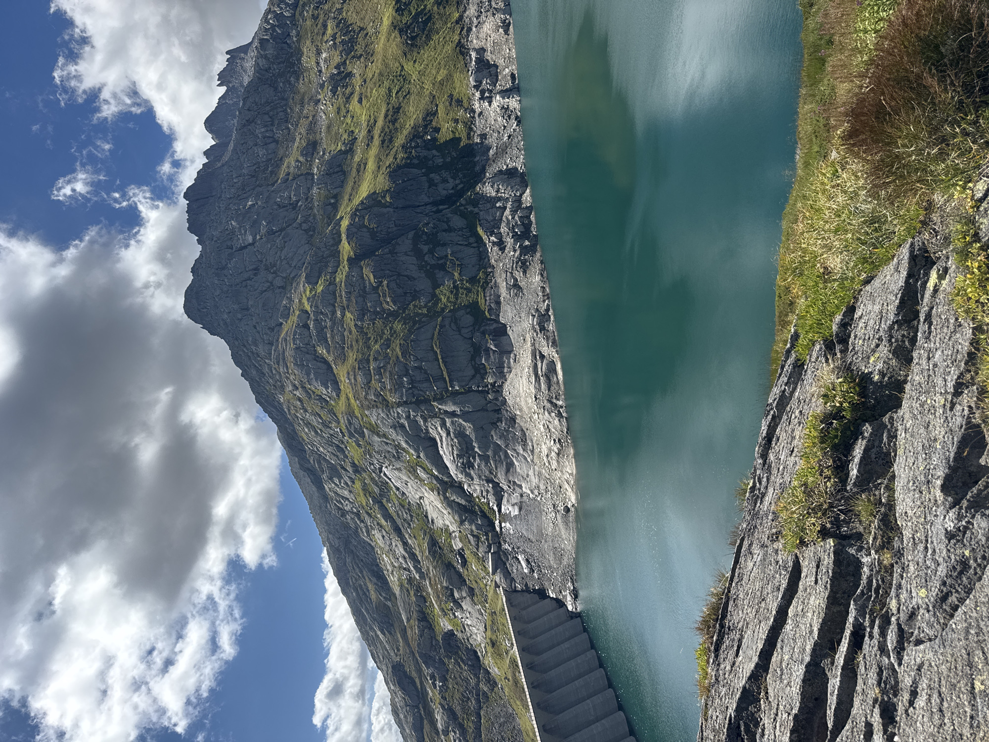 The image depicts a wide shot of the Grimsel Dam in Airolo, Switzerland, on a clear day. The dam, constructed from concrete, forms the left boundary of the scene, with its tiered structure clearly visible. Beyond the dam, a vast expanse of calm, turquoise water stretches across the frame. The background is dominated by a majestic, rugged mountain range, its rocky slopes partially covered in patches of green vegetation. The sky above is a vibrant blue, dotted with scattered white clouds. The foreground consists of a rocky, uneven terrain with sparse green grass and vegetation, suggesting a natural, mountainous environment. There are no people or animals visible in the image.