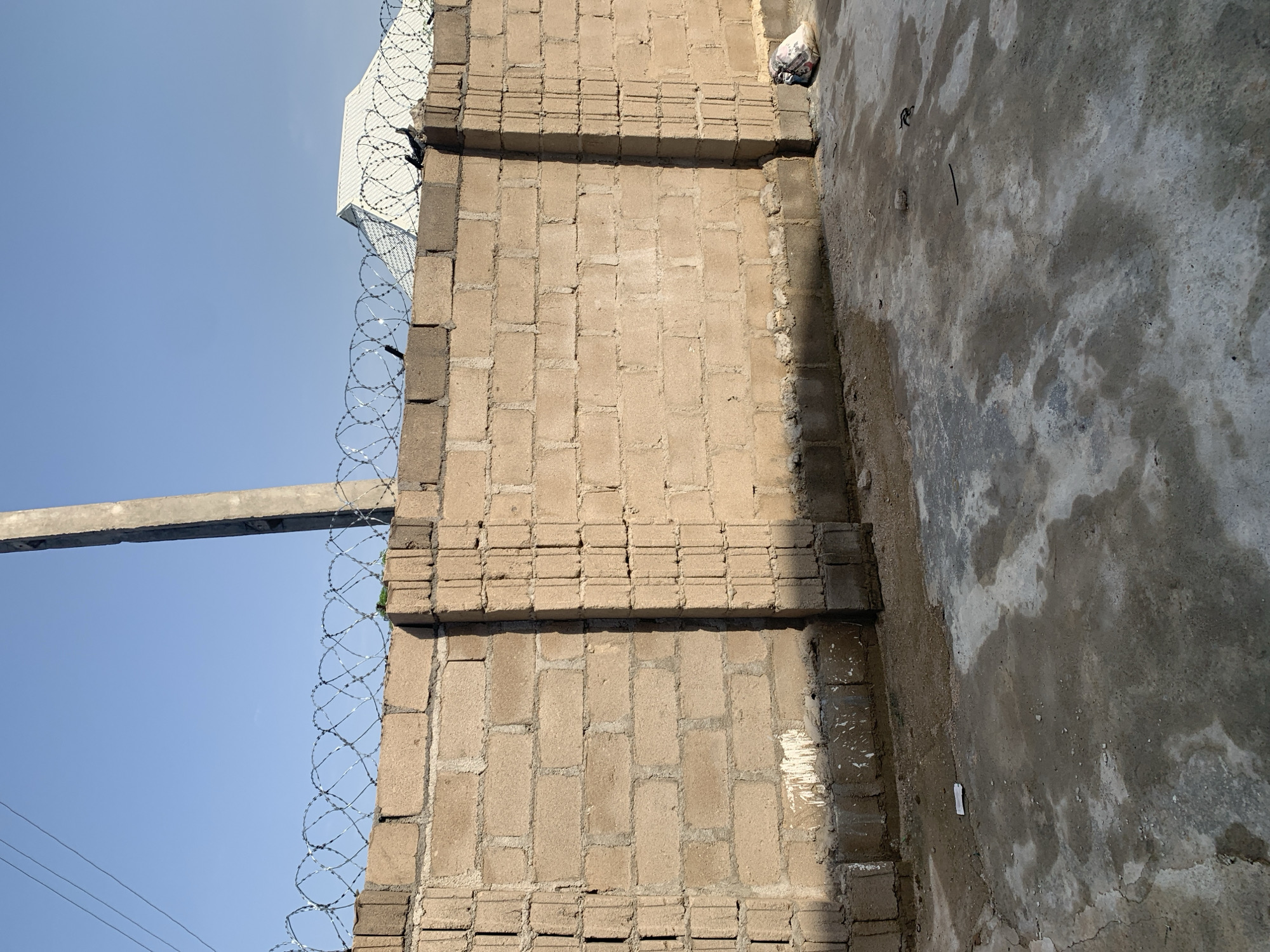 An outdoor, eye-level, full shot shows a concrete block wall topped with coiled barbed wire under a clear blue sky. The wall is a light tan color and is constructed from rectangular blocks laid in a staggered pattern. Vertical concrete pillars, also made of blocks, are spaced along the wall. The ground in front of the wall is a mix of concrete and dirt, with some areas appearing wet. A single, small white object, possibly a piece of paper, is visible on the ground in the lower left quadrant. In the lower right quadrant, a white bag filled with dark contents rests on the ground. The background shows the white, angled roof of a building and a tall, grey utility pole.