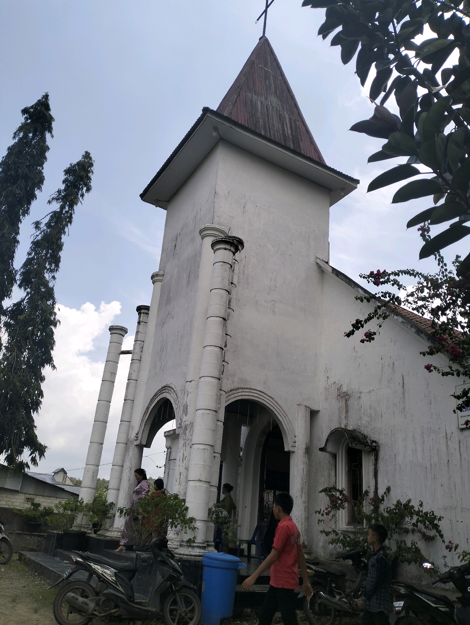 The image depicts a white, two-story building with a prominent steeple, suggesting it is a church. The building has arched doorways and windows. Several people are visible, including two women standing on steps, a man walking by, and two young boys in the foreground. The exterior is weathered and shows some signs of age. The surrounding environment includes trees, foliage with pink flowers, two motorcycles parked in front, and a blue trash can. In the background, there are other buildings and a cloudy sky. The scene is identified as being in the city of Ndraso Hilisimaetano, Indonesia.