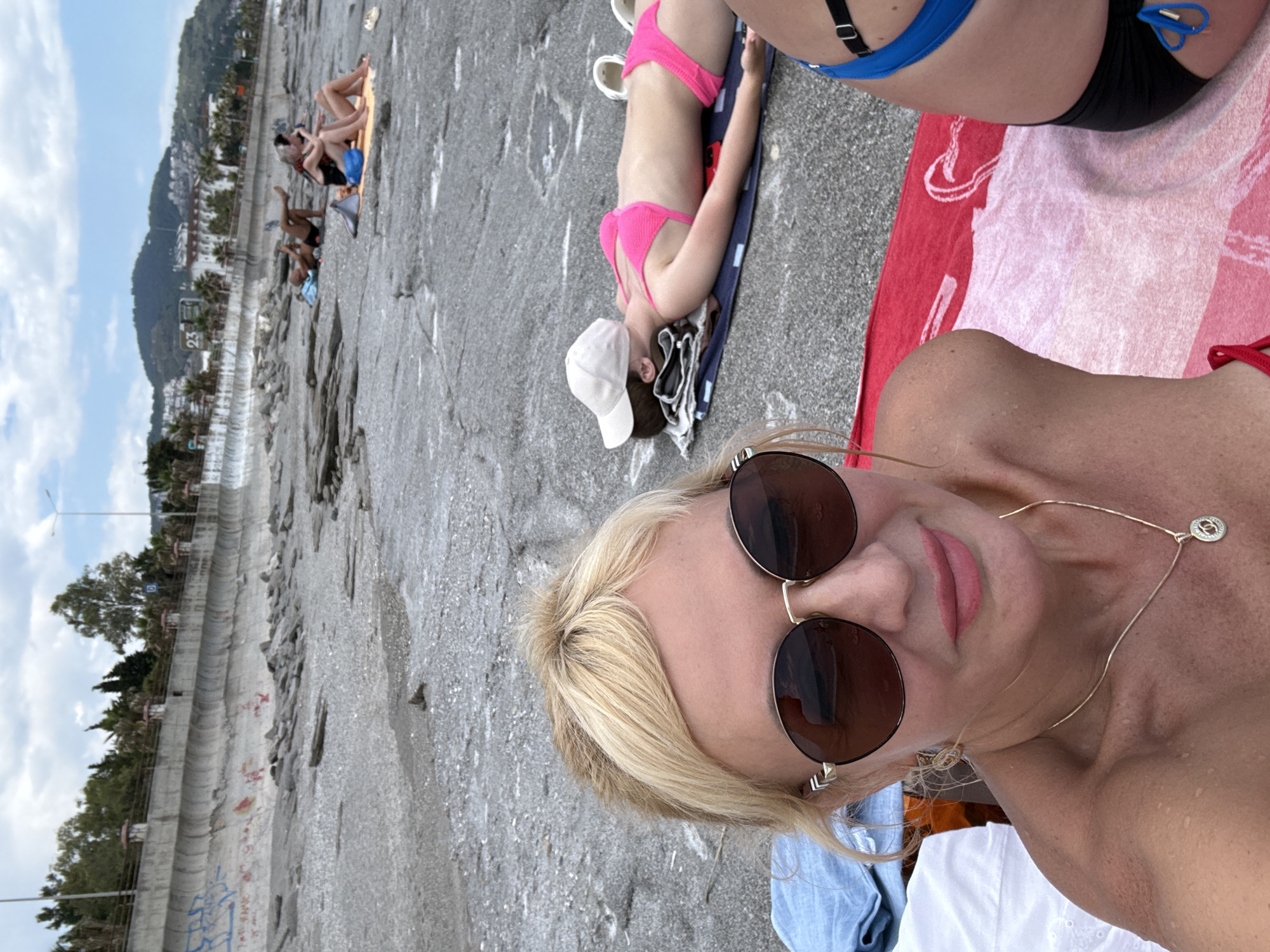The image shows a woman taking a selfie on a stone beach in an unknown city. She is wearing sunglasses and a necklace. Behind her, there is a woman in a pink bikini lying on a towel. There are other people relaxing on the beach in the background. The beach is adjacent to a waterfront structure and trees.