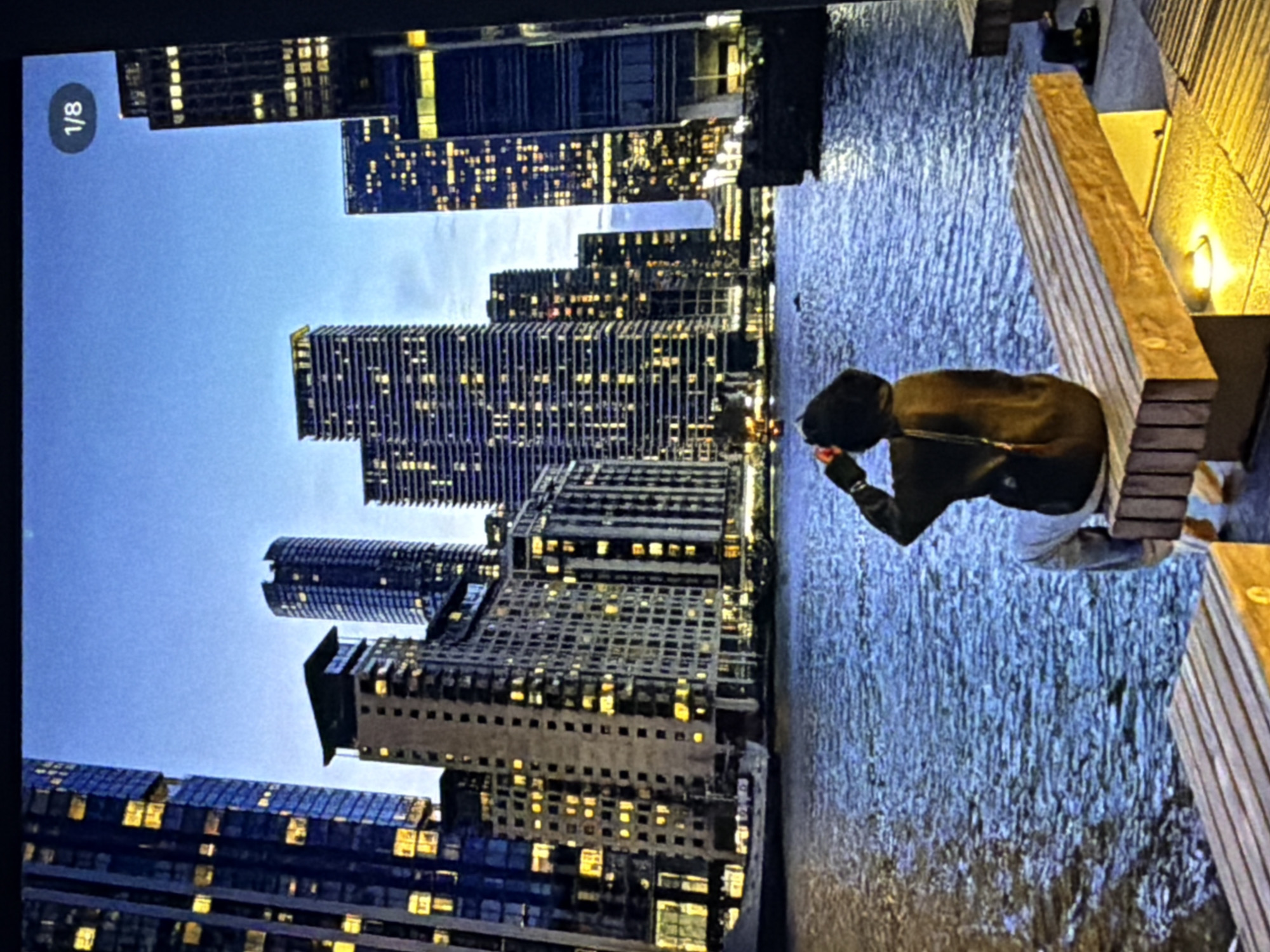 The scene depicts a cityscape at twilight or dawn in Maidstone, United Kingdom, viewed across a body of water. In the immediate foreground, a person is seated on a modern wooden slatted bench, facing away from the camera and looking towards the distant buildings. The individual wears a dark hooded top or jacket and light-colored trousers; their right arm is raised to the side of their head. The bench features integrated lighting at its base, emitting a warm yellow glow onto the paved ground. Another similar bench is partially visible in the lower-left foreground. The midground is occupied by a wide expanse of water, exhibiting numerous ripples on its surface, reflecting the ambient light and city lights. Across the water, a dense cluster of tall, modern high-rise buildings forms a prominent urban skyline. These buildings are predominantly dark-toned, with numerous windows, many of which are illuminated with warm yellow lights. Architectural styles vary, including multi-story glass-fronted towers on the left, rectangular structures with patterned facades in the center, and a distinctive slender, cylindrical or oval-shaped building towards the right of the center. On the far right, further tall, dark buildings with grids of lit windows are visible. A boat or ship is docked near the buildings on the left side of the water. The sky above the cityscape is a clear, deep blue, transitioning from a slightly lighter hue near the horizon.