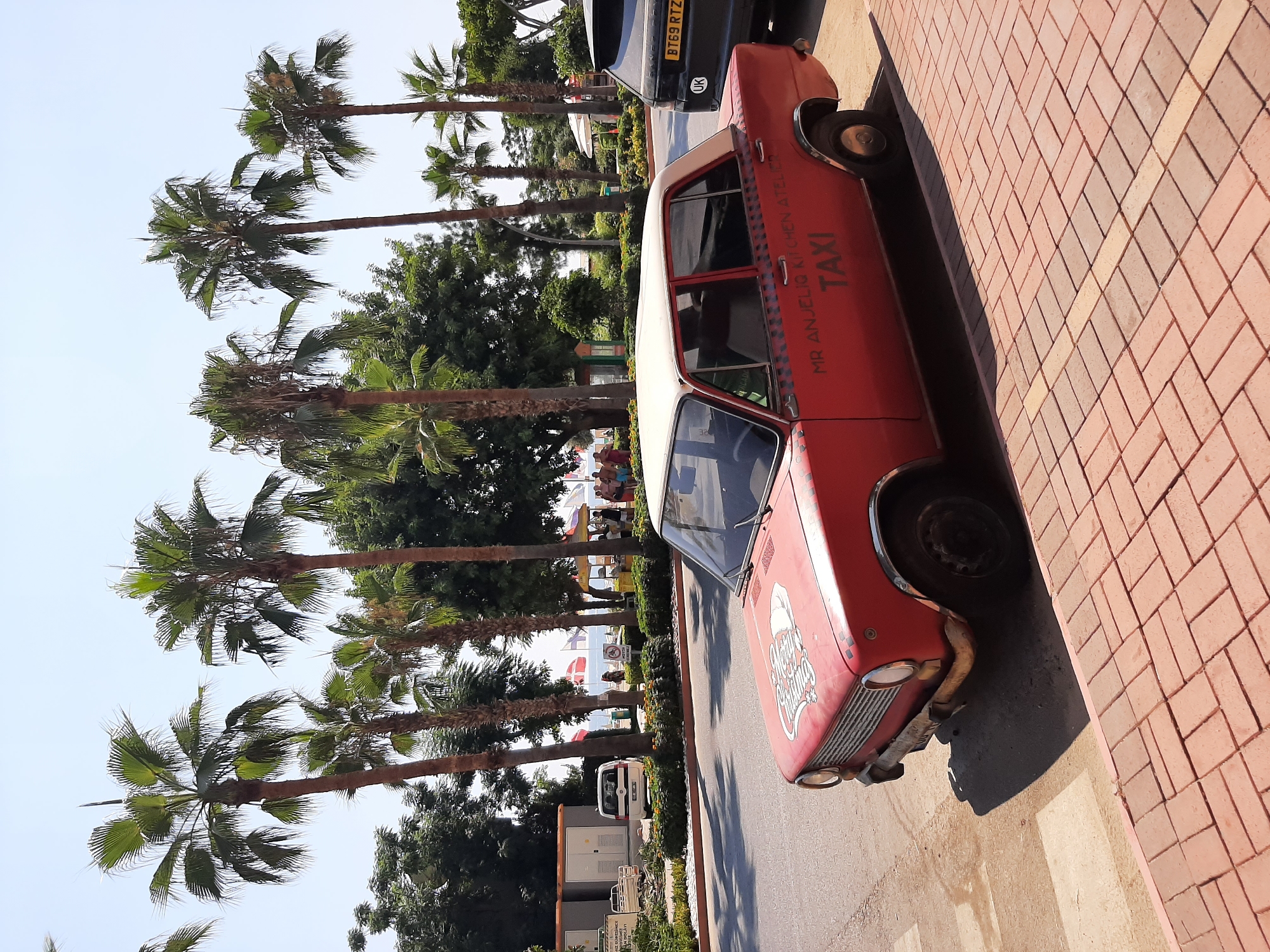 In Alanya, Turkey, a vintage red car, possibly a Lada, is parked on a brick-patterned sidewalk next to a street. The car has a white roof and text on its side reading "MR ANJELIQ KITCHEN ATELIER" and "TAXI." A black SUV with a UK license plate (BT69 RTZ) is parked behind it. In the background, several palm trees line the street, and beyond them, people are visible on a beach with umbrellas and flags, suggesting a coastal location. The sky is clear and blue. The scene appears to be a sunny day in a tourist area.