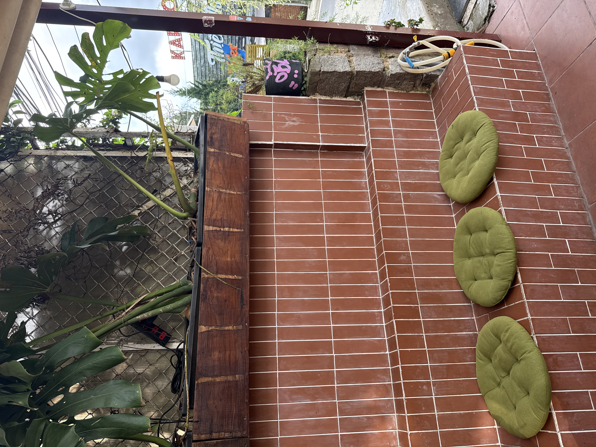 An outdoor patio space, possibly at a cafe or residence, is depicted with a focus on its seating and plant elements. No people are visible.
The scene is dominated by reddish-brown rectangular tiles with white grout, forming tiered surfaces or steps that define the layout. Three circular, tufted green cushions are laid out on these tiled steps, inviting seating. To the left, a robust wooden planter box or barrier runs horizontally, acting as a support for lush vegetation. Behind this, a chain-link fence is visible, with more greenery and vines growing through it.
A large Monstera plant with characteristic perforated leaves, some showing signs of yellowing or spotting, extends prominently from the left side of the frame. Other smaller green plants and foliage are scattered throughout, contributing to a vibrant, natural feel.
In the upper background, on a raised tiled ledge, sits a black plant pot adorned with pink graffiti that appears to spell "MOO." Nearby, a coiled white garden hose with yellow and blue connectors is placed. Overhead, a single white lightbulb hangs from a wire. Various other cables and wires are visible, including one connected to a dark object with red indicator lights secured to the chain-link fence. Small decorative stickers are affixed to a metal beam spanning the top of the area.
The time of day is daytime, under an overcast or cloudy sky, providing soft, diffused light. The setting suggests a casual, laid-back outdoor environment, characteristic of places like Da Lat, Vietnam. Partial text "KAA" is visible in red on a white sign in the background, along with "vie" on another sign further back.