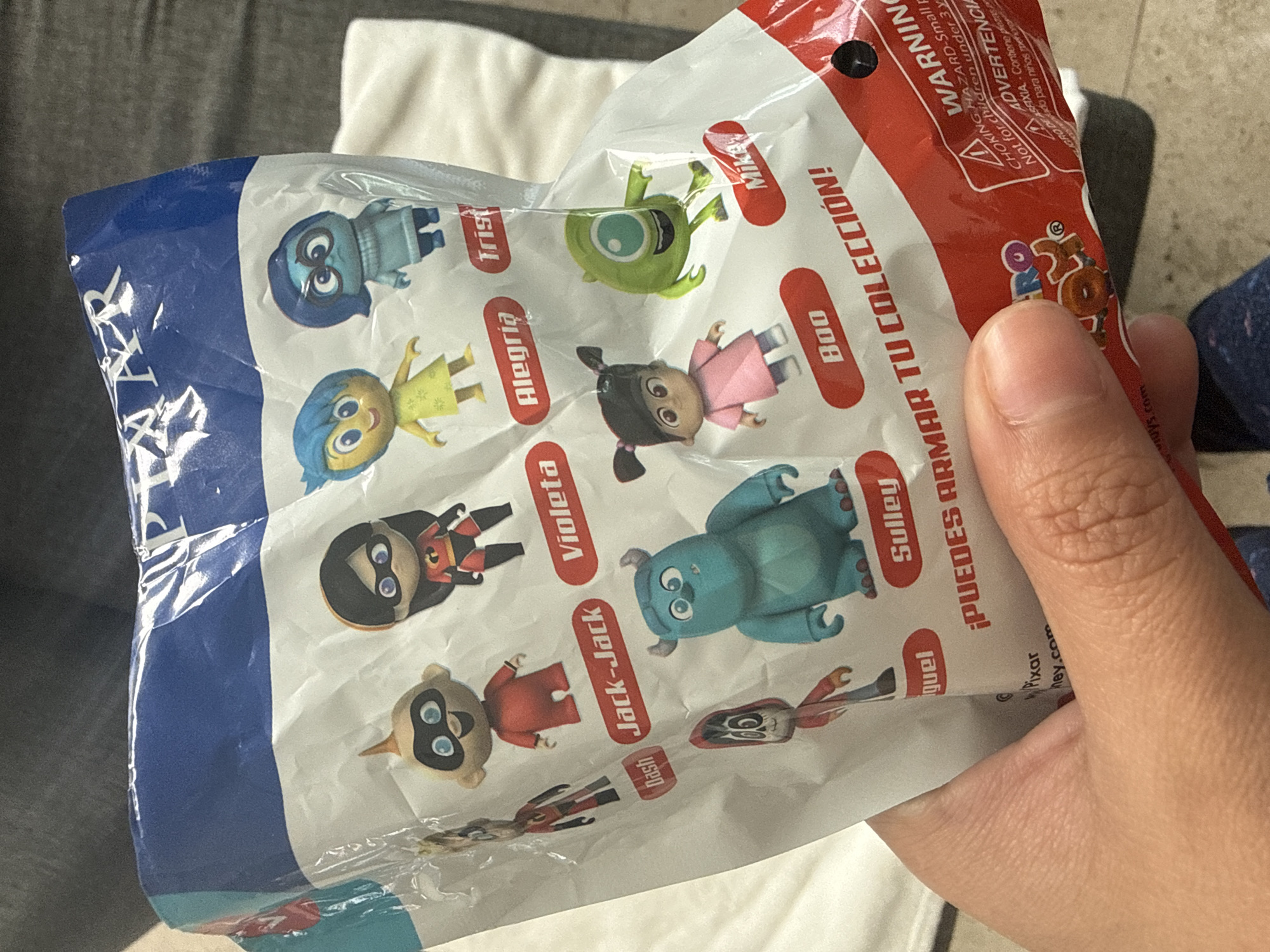 The image shows a person's hand holding a sealed packet of Disney Pixar figurine collectibles. The packet displays images of various characters, including Sadness, Joy, Mike, Boo, Sulley, Miguel, Dash, Jack-Jack, and Violeta, with their names written in Spanish (Trist, Alegria, Mike, Boo, Sulley, Guel, etc.) "¡PUEDES ARMAR TU COLECCIÓN!" is printed on the packet. A warning label in English and Spanish is visible near the top of the image. The background is blurry and shows a gray sofa, a white pillow, and blue and white fabric. The image does not provide specific details about the location being San Bernardino Tlaxcalancingo, Mexico.