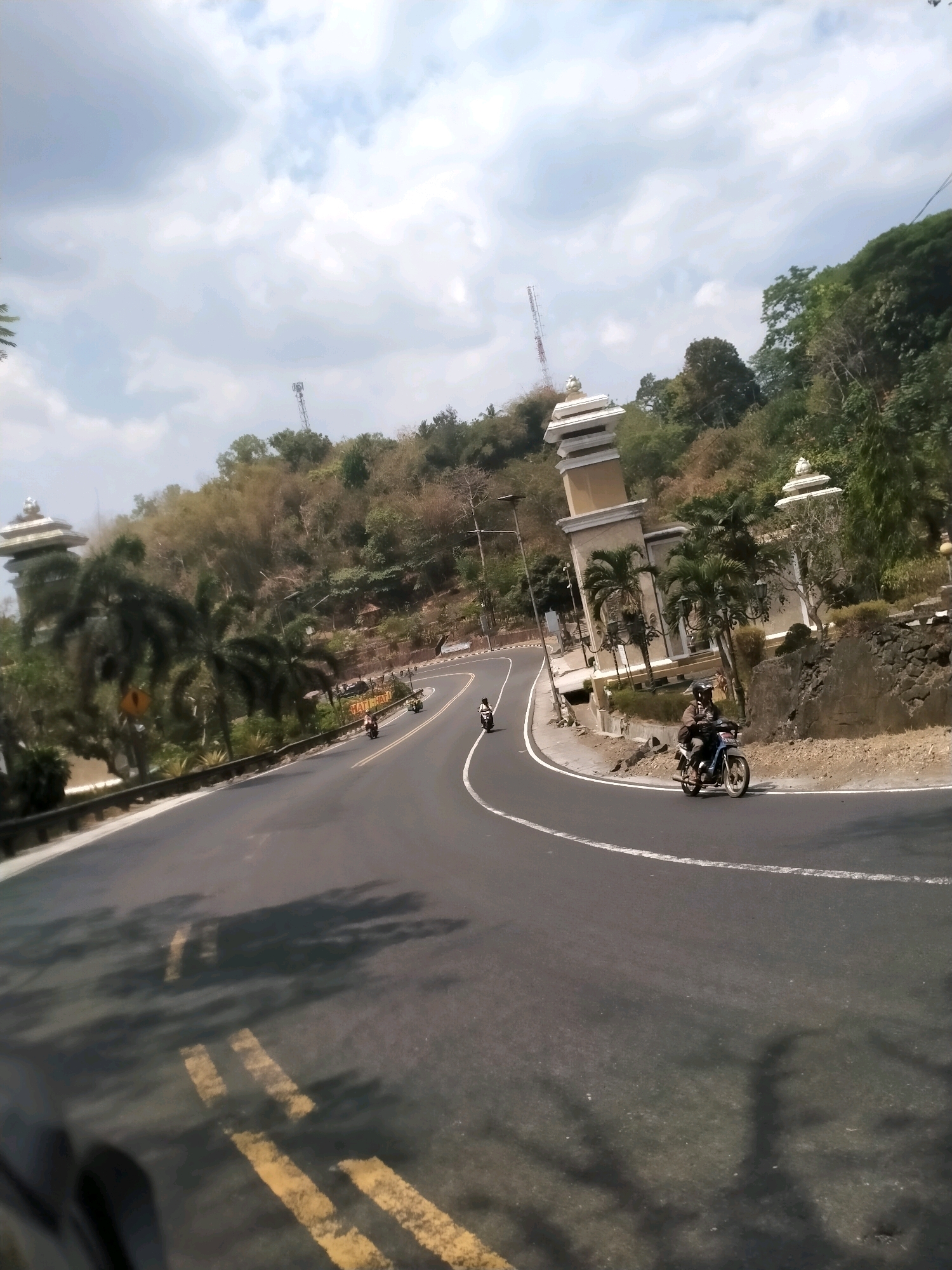 The image depicts a winding asphalt road with yellow lane markings, likely in Patuk, Indonesia, given the context. The road curves to the left, ascending a hillside. On the right side of the road, a rocky embankment is visible, topped with vegetation and a distinctive white and tan architectural structure with a tiered roof, resembling a watchtower or pavilion. Palm trees are scattered along the roadside. Several motorcycles are on the road. In the foreground and mid-ground, two motorcycles are clearly visible, each with a rider wearing a helmet. One motorcycle, on the right, is blue and white. Another motorcycle, further up the road on the left, is also visible. The sky is partly cloudy. The overall scene suggests a scenic route with some developed infrastructure.