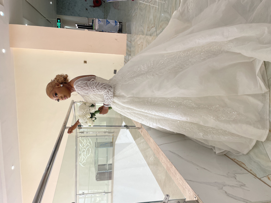 The image portrays a female subject in a formal white wedding gown, situated indoors within a multi-level building in Barakin Lerawi, Nigeria. The woman stands facing slightly left, her right hand resting on a silver-colored metal handrail, which tops a glass and marble-patterned balustrade. She is holding a bouquet of white flowers in her left hand. Her blonde hair is styled in an updo, and she wears square earrings. The gown features long, sheer, beaded sleeves, a voluminous skirt, and a trailing train with sparkling embellishments. The floor is tiled with light-colored, possibly marble, panels. The walls are light beige, transitioning to a lighter color in the background right. Visible in the background are a green exit sign, two cylindrical wheeled bins (one silver, one dark), and a red fire hose reel mounted on the wall. The lighting is bright and even.