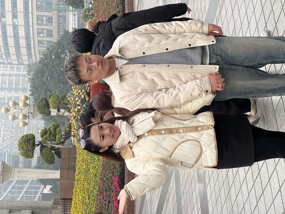 A couple poses for a photo amidst city scenery. The woman, wearing a quilted white jacket and dark skirt, smiles at the camera while gesturing with her hand. Her partner, beside her, is dressed in jeans and a similar white jacket, with glasses. They stand among flowers and trimmed trees with city buildings in the background. Other people are also around them.