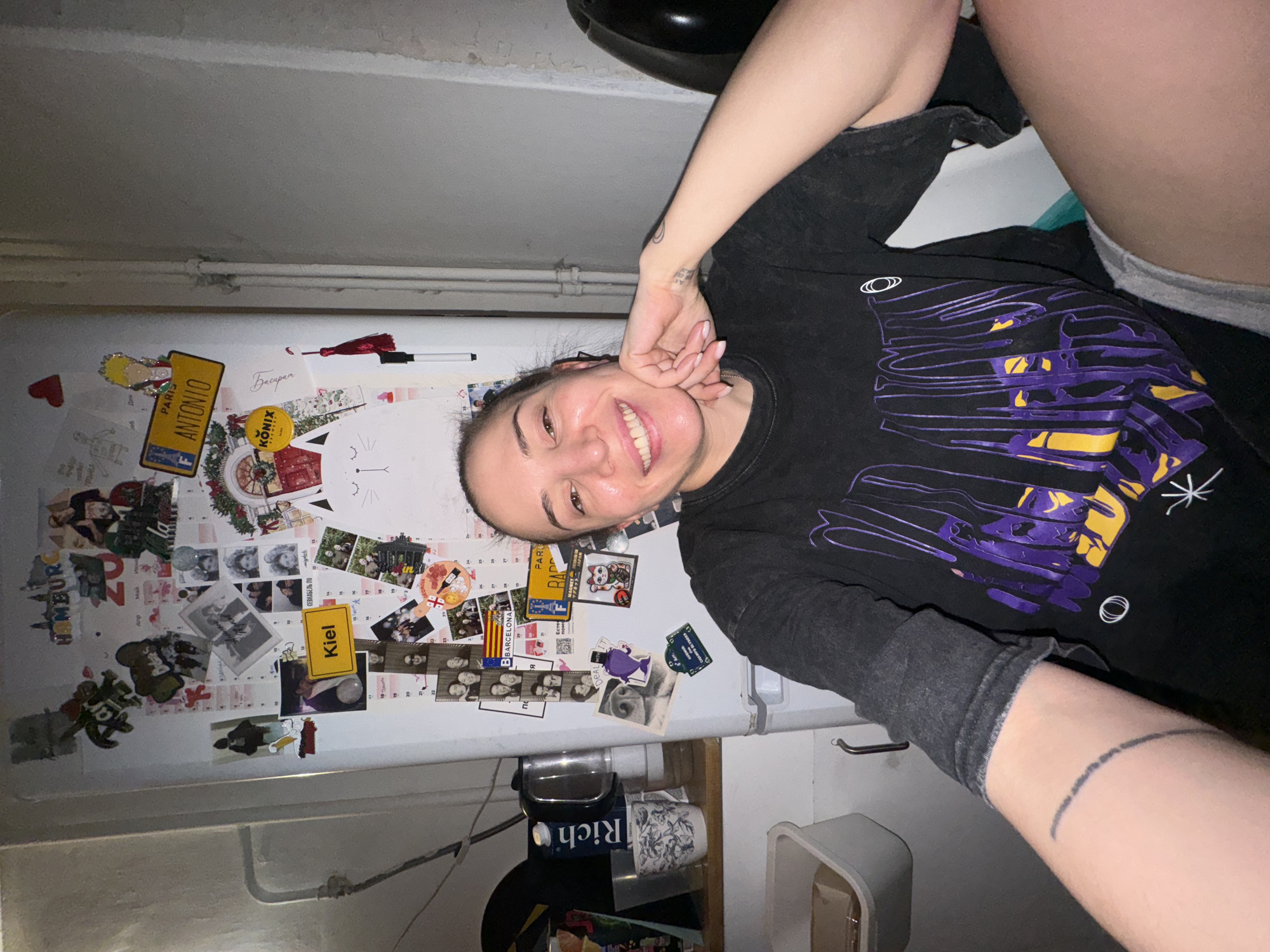 A selfie of a smiling woman with dark hair pulled back is taken in front of a refrigerator door covered with magnets, photos, and memorabilia. The woman is wearing a dark, graphic t-shirt and grey shorts. Her left hand is near her face and the right arm is visible, featuring a tattoo of a line on her forearm. The refrigerator door is covered in various items, including license plate magnets labeled "Paris Antonio," "Kiel," and "Homburg." Other items visible on the refrigerator include a "Barcelona" magnet, calendar pages, photos of people, and a drawing of a cat face. The background reveals a cluttered room, including a sink, a black trash can, and other containers. There is no evidence to suggest this image was taken in Moscow, Russia.