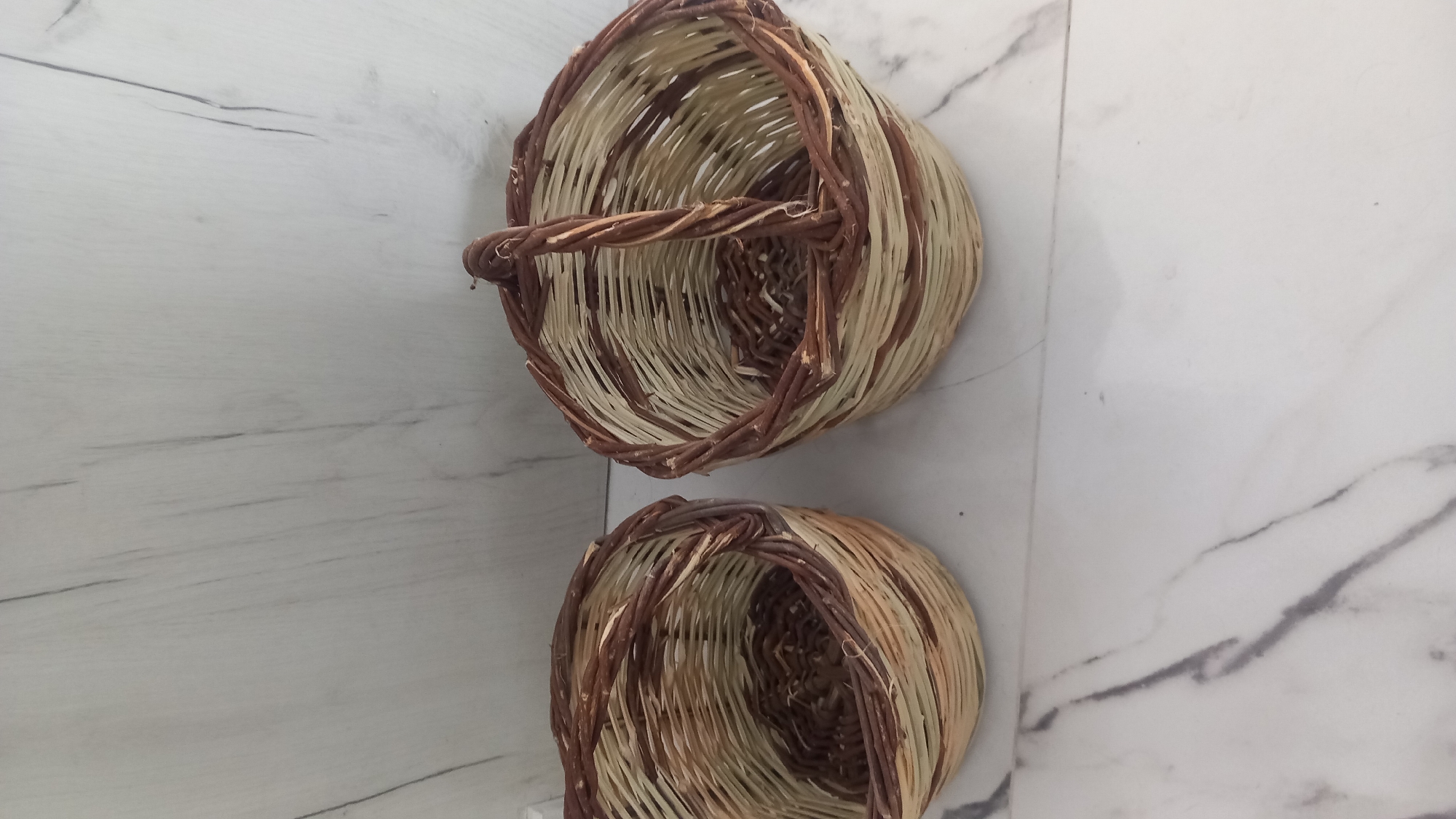 Two woven wicker baskets are placed on a marble-patterned surface in front of a light-colored, wood-patterned wall. The basket on the left is wider and shorter with an irregular opening. The basket on the right is taller and narrower, and it has a curved handle made of twisted wicker. Both baskets are constructed from light-colored and dark brown wicker. The scene appears to be indoors, possibly in a home or a craft studio, in a city like Άγιος Νεκτάριος, Greece. There are no people visible in the image.