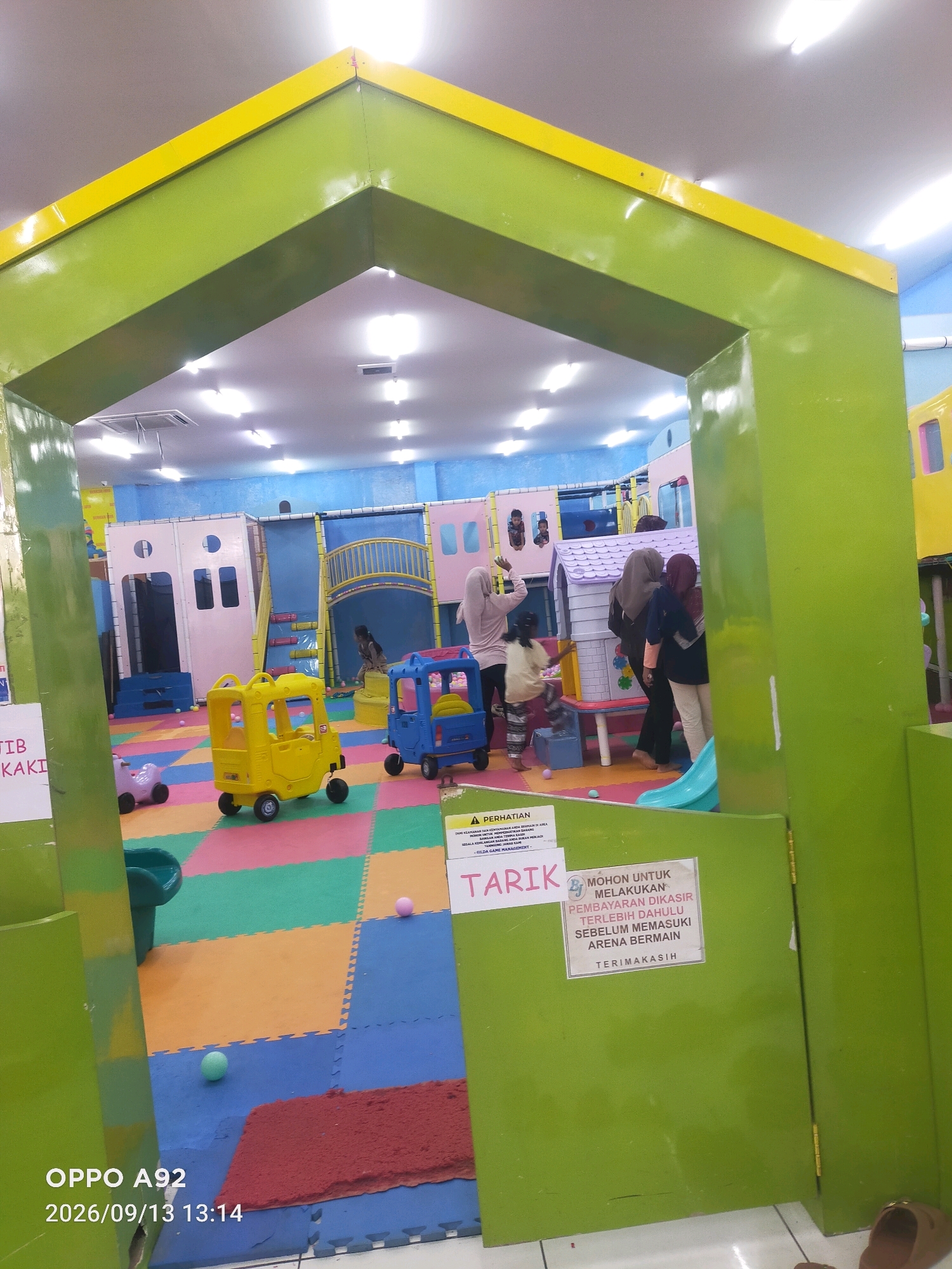 The image captures a vibrant indoor playground in Kubangkangkung, Indonesia, with children and adults present. The playground features colorful foam floor mats in various shades of blue, green, yellow, and pink, forming a cheerful and safe play area. Dominating the foreground is a bright yellow toy car, while a blue toy car is also visible. In the background, a multi-level play structure with pink and blue sections and a yellow slide can be seen, suggesting a space designed for active play and exploration.
Several individuals are interacting within the playground. Two adult women, one wearing a light pink hijab and the other a dark headscarf, are observed near the play structure. A young girl in a white shirt and patterned pants is actively engaged in play, reaching towards the structure. Another child, partially obscured, is also near the play area. The lighting in the space appears to be artificial, emanating from ceiling lights.
Two signs are prominently displayed. One, a yellow and white sign with a red triangle warning symbol, reads "PERHATIAN" (Attention) and provides safety instructions in Indonesian. Another white sign with a red border and text in Indonesian states "MOHON UNTUK MELAKUKAN PEMBAYARAN DI KASIR TERLEBIH DAHULU SEBELUM MEMASUKI ARENA BERMAIN TERIMAKASIH" (Please make payment at the cashier first before entering the play arena, Thank You). The overall environment suggests a recreational facility designed for children's entertainment and safety.