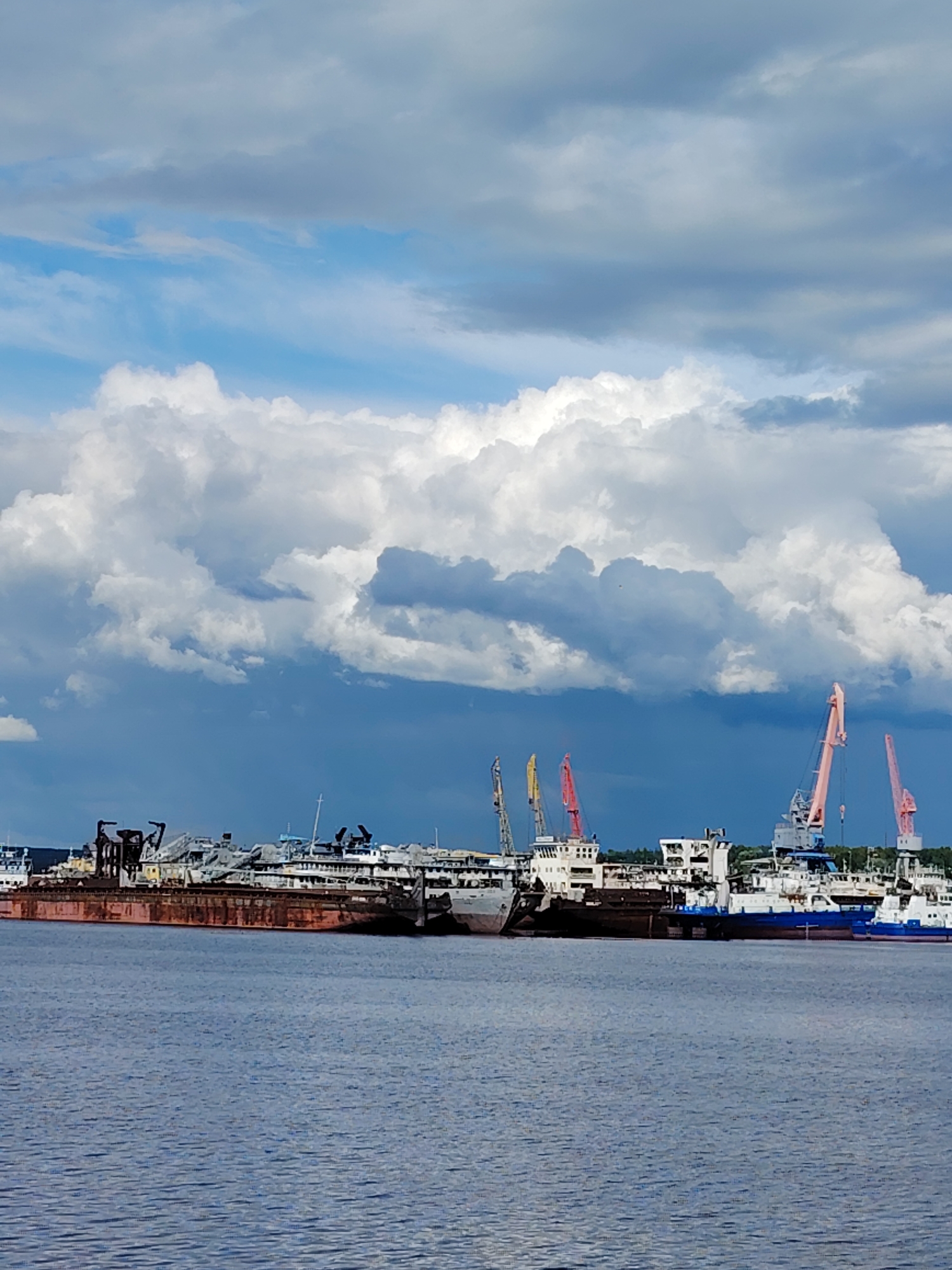 The image depicts a waterfront scene in the city of Chaikovsky, Russia, characterized by a mix of industrial elements and natural features.
The foreground is dominated by a body of water with a visible surface texture suggestive of wind-driven ripples. In the middle ground, a series of ships and barges are docked along the waterfront. These vessels vary in size, color, and apparent condition, with some showing signs of rust and disrepair. Cranes of different colors (pink, yellow, red) are positioned behind the ships, adding to the industrial aesthetic.
The background is filled with a dramatic sky featuring puffy cumulus clouds, some bright white and others darker, indicating varying degrees of sunlight and perhaps an approaching storm. A thin line of trees or vegetation is visible on the horizon, behind the ships and cranes.
Overall, the scene conveys a working industrial waterfront environment with the juxtaposition of natural and man-made elements.