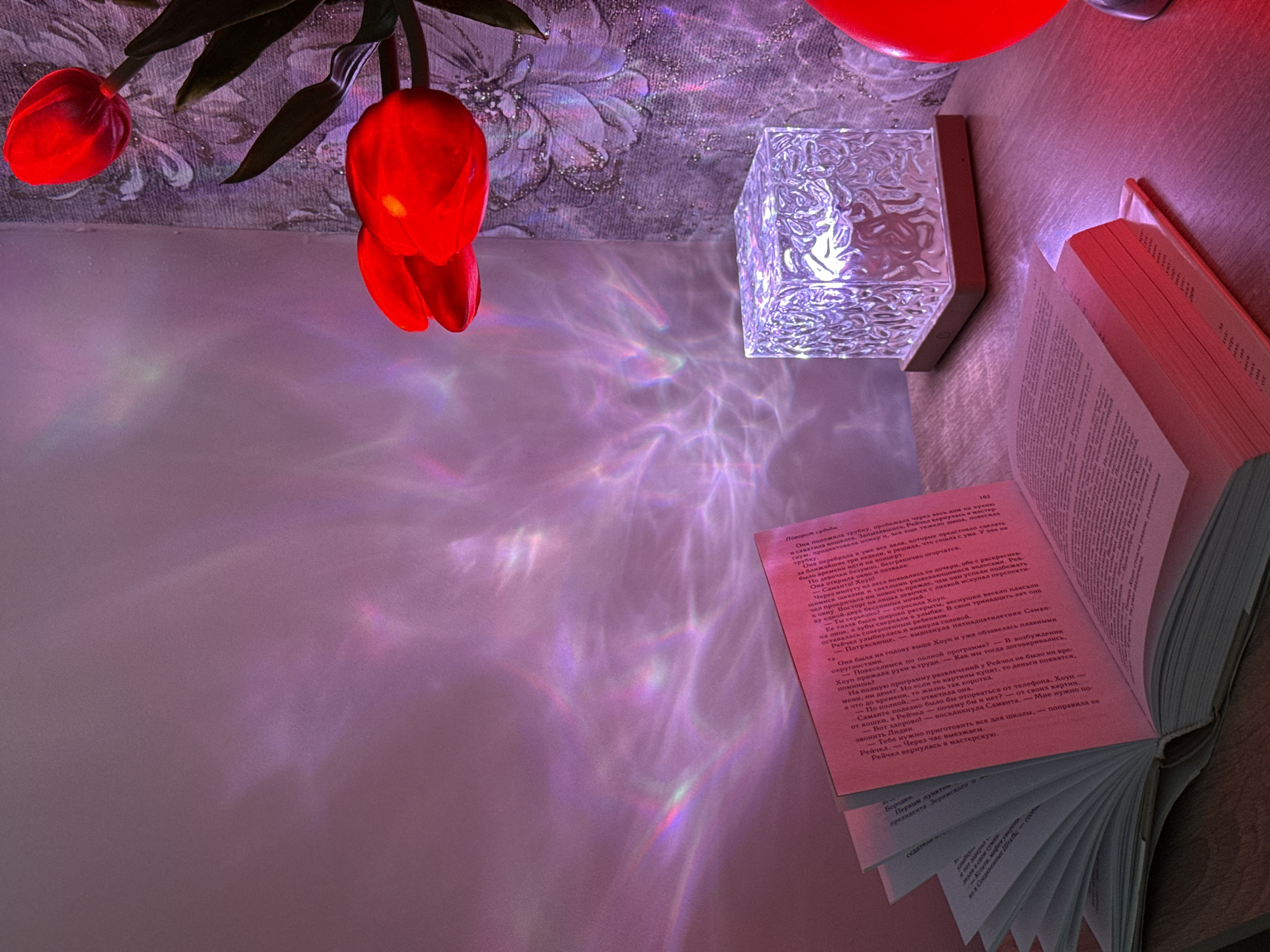 A high-angle shot captures a scene of books, a lamp, and flowers on a surface. The book, open with text visible, is prominently placed. Next to it, a cube-shaped lamp diffuses light, creating a pattern of reflections on the surface. Red tulips are partially visible at the top left of the frame.