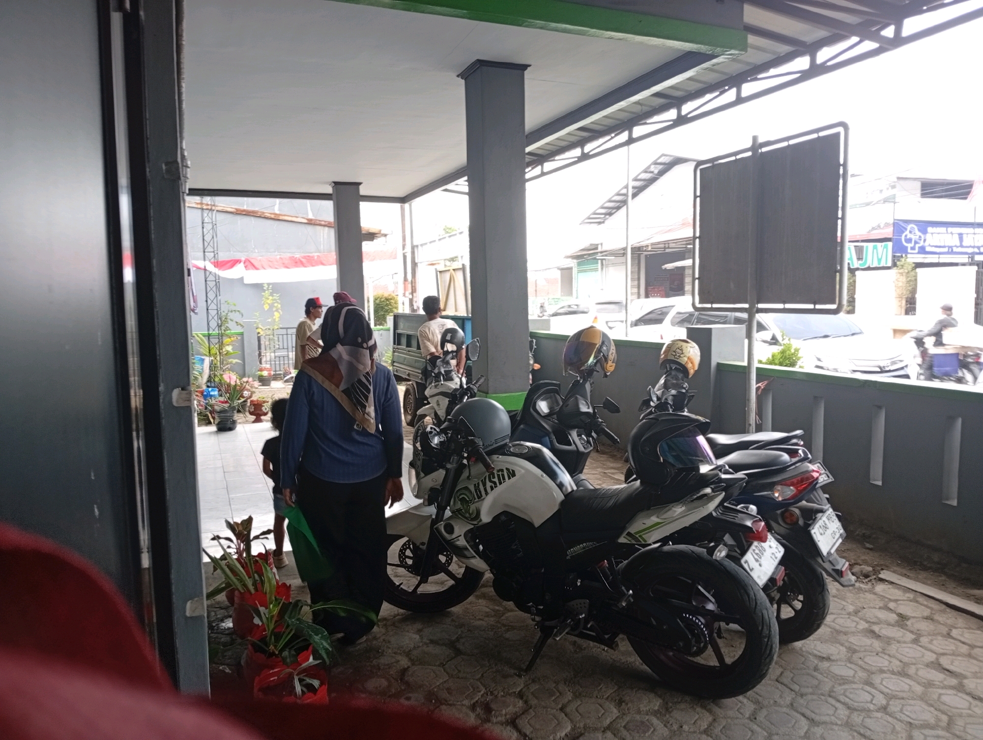 The image shows an outdoor area with a covered overhang supported by columns. Several motorcycles are parked under the overhang, with one white and green motorcycle prominently displayed in the foreground, featuring the text "BYSON" on its fuel tank and a helmet on its handlebars. To the right of this motorcycle, two other dark-colored motorcycles are parked, one with a helmet on its seat. In the background, several people are visible, including a woman wearing a blue hijab and dark clothing standing next to the white motorcycle, and a child wearing a dark shirt and shorts standing behind her. Further back, other individuals are seen in lighter clothing, and some are near what appears to be a delivery truck or service vehicle with a white tarp. A large blank sign is mounted on a post in the middle ground. The ground is paved with hexagonal tiles. The overall environment suggests a commercial or service area in a town or city, likely in a tropical climate given the greenery and open-air setting. The image appears to be taken during daylight hours, with bright, overcast skies. The setting suggests a location in Tasikmalaya, Indonesia, consistent with the presence of motorcycles, typical architecture, and outdoor commercial spaces often found in Indonesian cities.