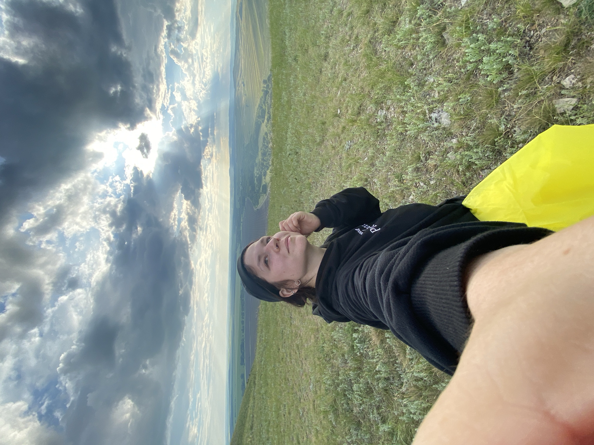 A young woman wearing a black hoodie and a black cap is taking a selfie on a grassy hill overlooking a vast rural landscape under a cloudy sky. The sun is breaking through the clouds, casting rays of light onto the scene. The woman is looking upwards and to her right, with her hand near her face. A bright yellow object, possibly a bag or piece of clothing, is visible on her lap. The location is the village of Абзаново, Russia.