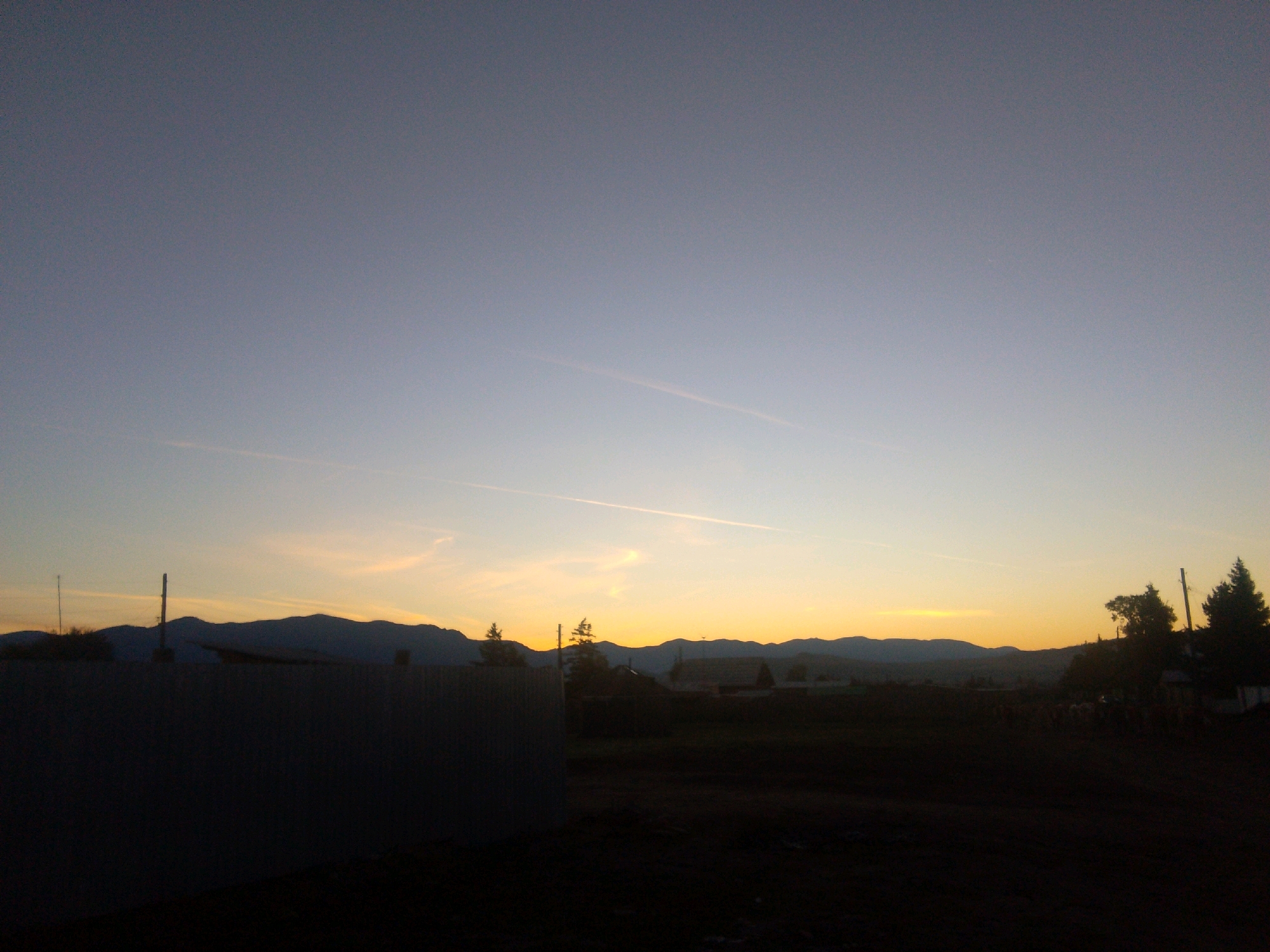 The image depicts a rural landscape at dusk, possibly in the city of Суш, Russia, given the context. A large, dark, textured wall or fence occupies the foreground on the left side. Behind this structure, the landscape transitions into dark, silhouetted trees and indistinct buildings against a backdrop of rolling hills or mountains. The sky is a gradient of soft blues and yellows, indicative of a sunset. Faint contrails are visible in the upper portion of the sky. The overall impression is one of a quiet, rural evening. There are no people or specific actions clearly visible in the frame. The lighting is low, creating deep shadows across the ground and obscuring finer details of the terrain and structures.