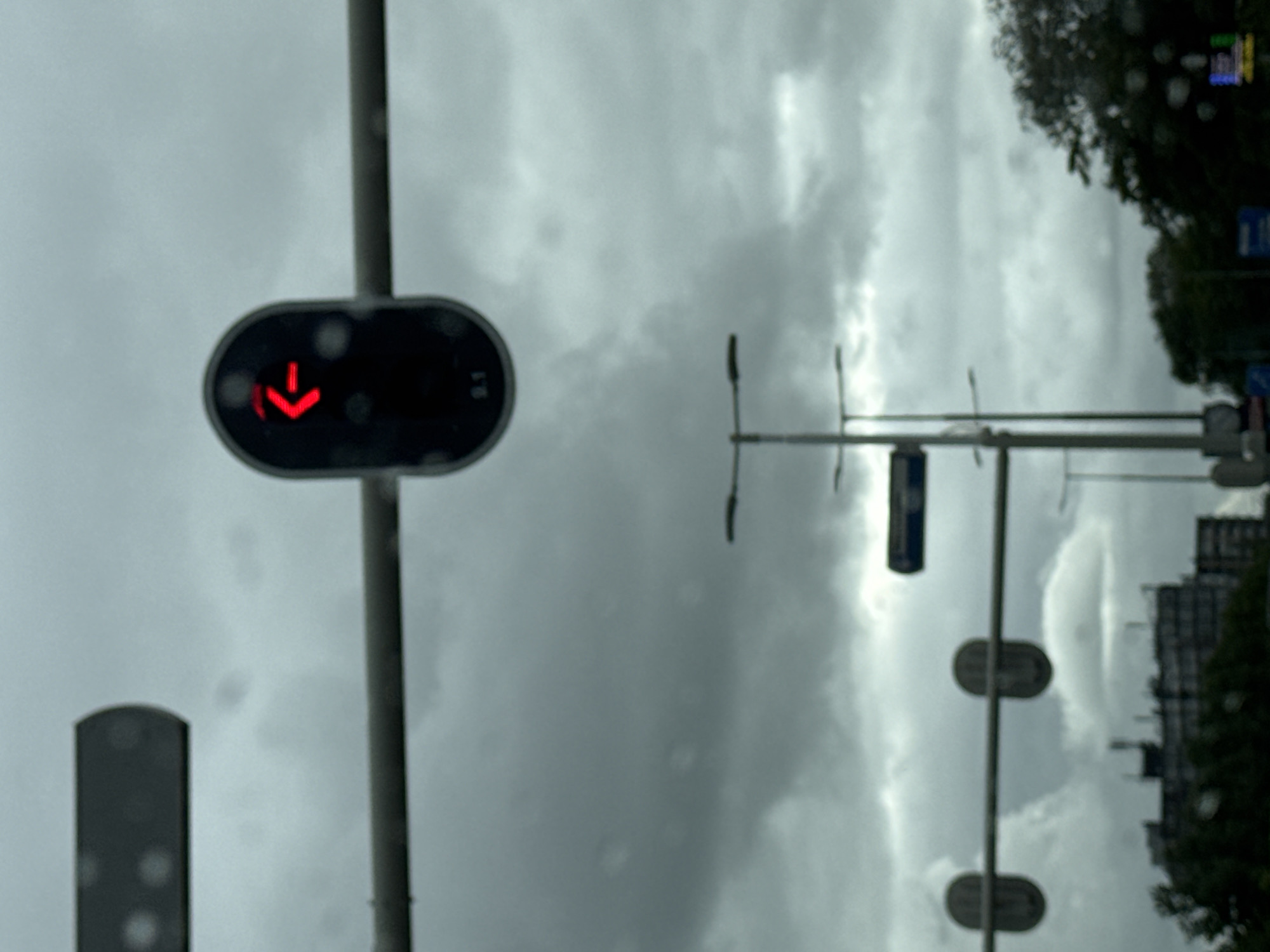 This is a photograph taken from a vehicle, likely during rainfall, as evidenced by water droplets on the windshield. The scene captures a traffic light with a red left-pointing arrow illuminated, indicating a restriction on turning left. The traffic light is suspended over a road, with other traffic lights and streetlights visible in the background. The sky is overcast with dark clouds, suggesting a gloomy or stormy weather condition. In the distance, buildings and trees can be seen. The composition and elements suggest an urban environment in Leeuwarden, Netherlands.