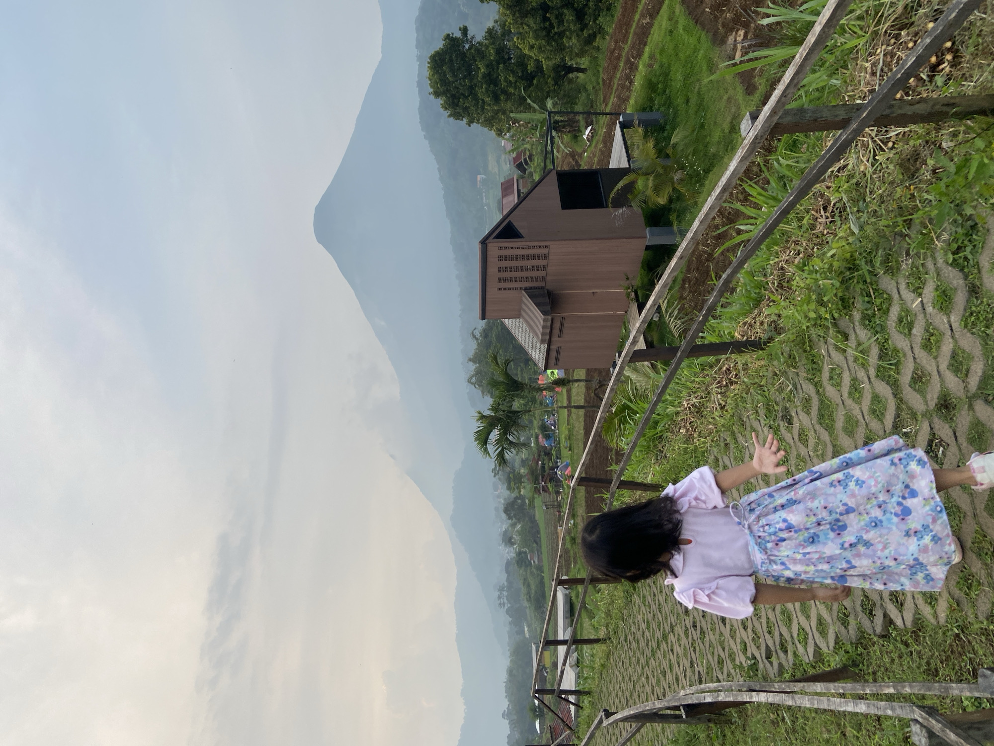 A girl stands on a stone path lined with a wooden fence, near a wooden building, with misty mountains in the background. The girl wears a light pink top and a blue floral skirt, with her back to the camera. The fence runs along the right side of the path, with green vegetation growing nearby. Beyond the fence, there is a brown wooden building with a unique design. In the distance, mountains are visible through a layer of mist, partially obscuring the landscape. Further beyond, several small tents dot the landscape in the area near Selotapak, Indonesia. The sky appears overcast and hazy.