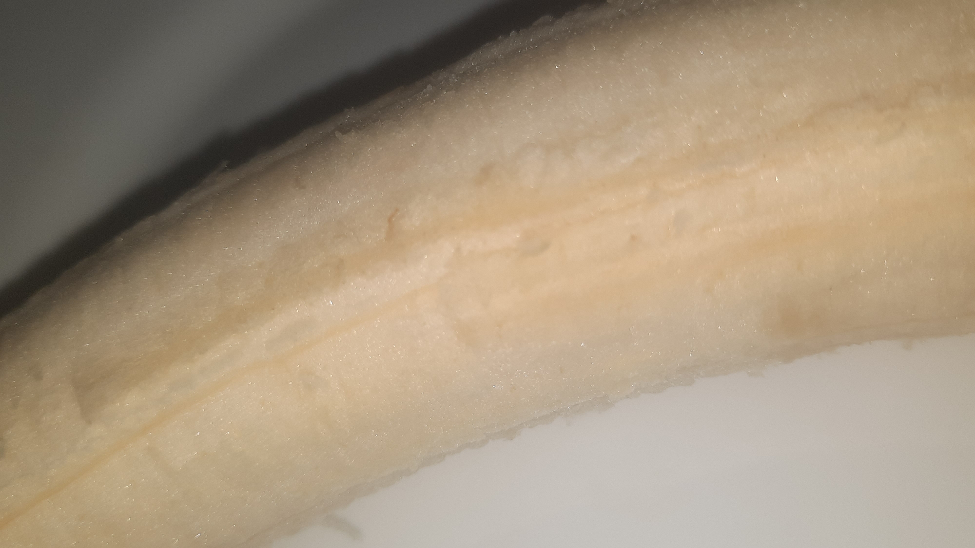 The image shows a close-up, detailed view of the interior of a peeled banana. The banana's flesh is a pale yellow, with visible fibrous strands running lengthwise along its surface. There are subtle darker yellow or light brown streaks and areas of discoloration, suggesting natural variations in the fruit. The texture appears soft and somewhat moist. The background is a plain, light-colored surface, likely a plate or countertop, with a gradient of light and shadow on the right side. There are no people or any indications of the city of Шклоў, Belarus present in the image.
