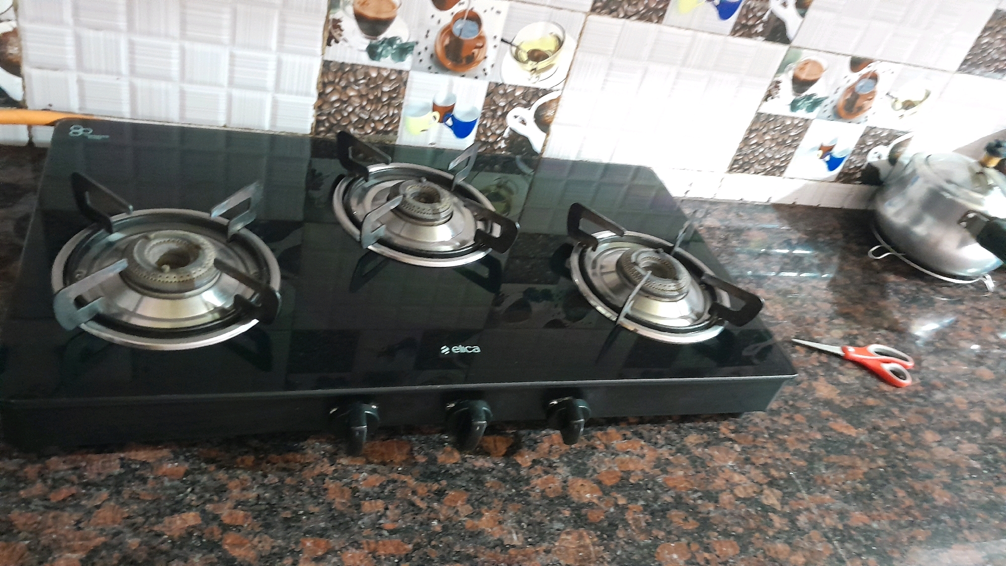 A three-burner black glass gas stove is positioned on a speckled brown granite countertop. The stove has a silver burner grate and a silver cooking grate above each burner. To the right of the stove, on the countertop, is a silver pressure cooker with a black handle and a small pair of red-handled scissors. The background features a tiled backsplash with a pattern of coffee cups and abstract designs. The scene is indoors, likely in a kitchen.