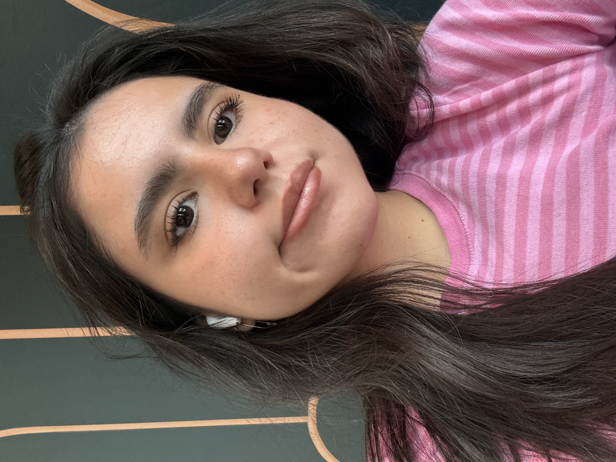 A young woman is seen in a close-up shot, wearing a pink and light pink striped shirt. She has dark hair and light skin with visible freckles. She has prominent dark eyebrows, brown eyes, and full lips. An earbud is visible in her ear. The background appears to be a dark wall with light brown vertical stripes. The location cannot be confirmed without additional information to be in the city of Austin, United States of America.