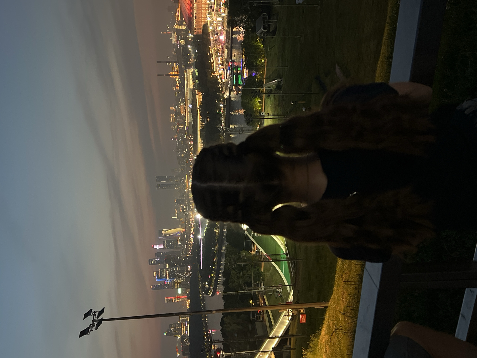 The image shows a young girl with long, braided brown hair pulled back into two pigtails, standing with her back to the camera and looking out at a cityscape at dusk. She is wearing a dark shirt. In the foreground, she is leaning on a railing. Behind her, a park area with green grass and trees is visible, as well as a ski jump structure and ski lifts. Beyond the park, the Moskva River flows, and boats with lights are on the water. In the distance, a dense collection of illuminated skyscrapers, characteristic of Moscow's Moscow City business district, dominates the skyline. The sky is a gradient of twilight colors, with soft clouds. The overall scene captures an urban panorama at the end of the day.