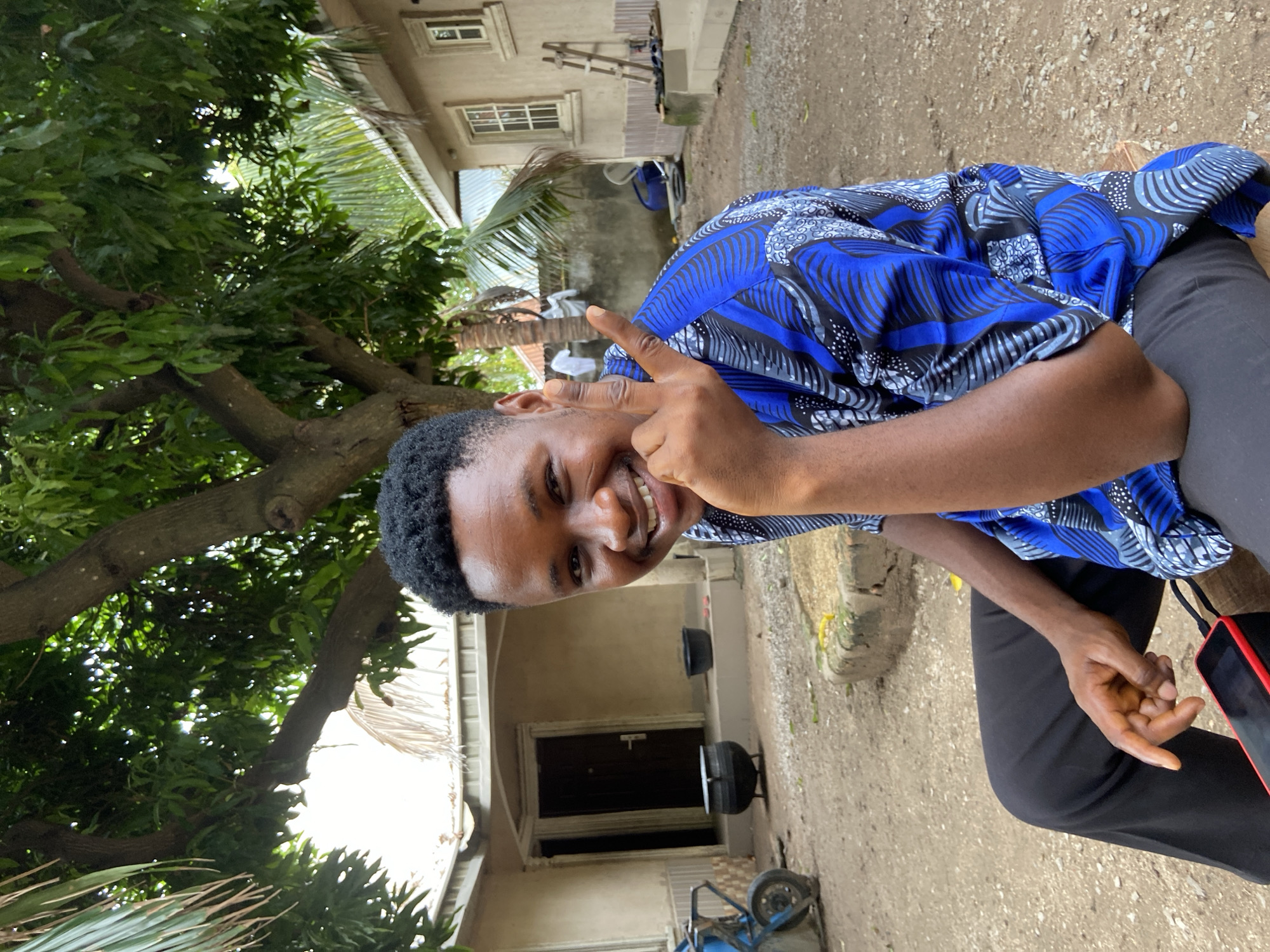 A young man sits outdoors in Mararaba, Nigeria, giving a peace sign. He wears a blue patterned shirt and black pants. A house stands behind him, and a tree is to his left. A blue handcart is in front of the house.