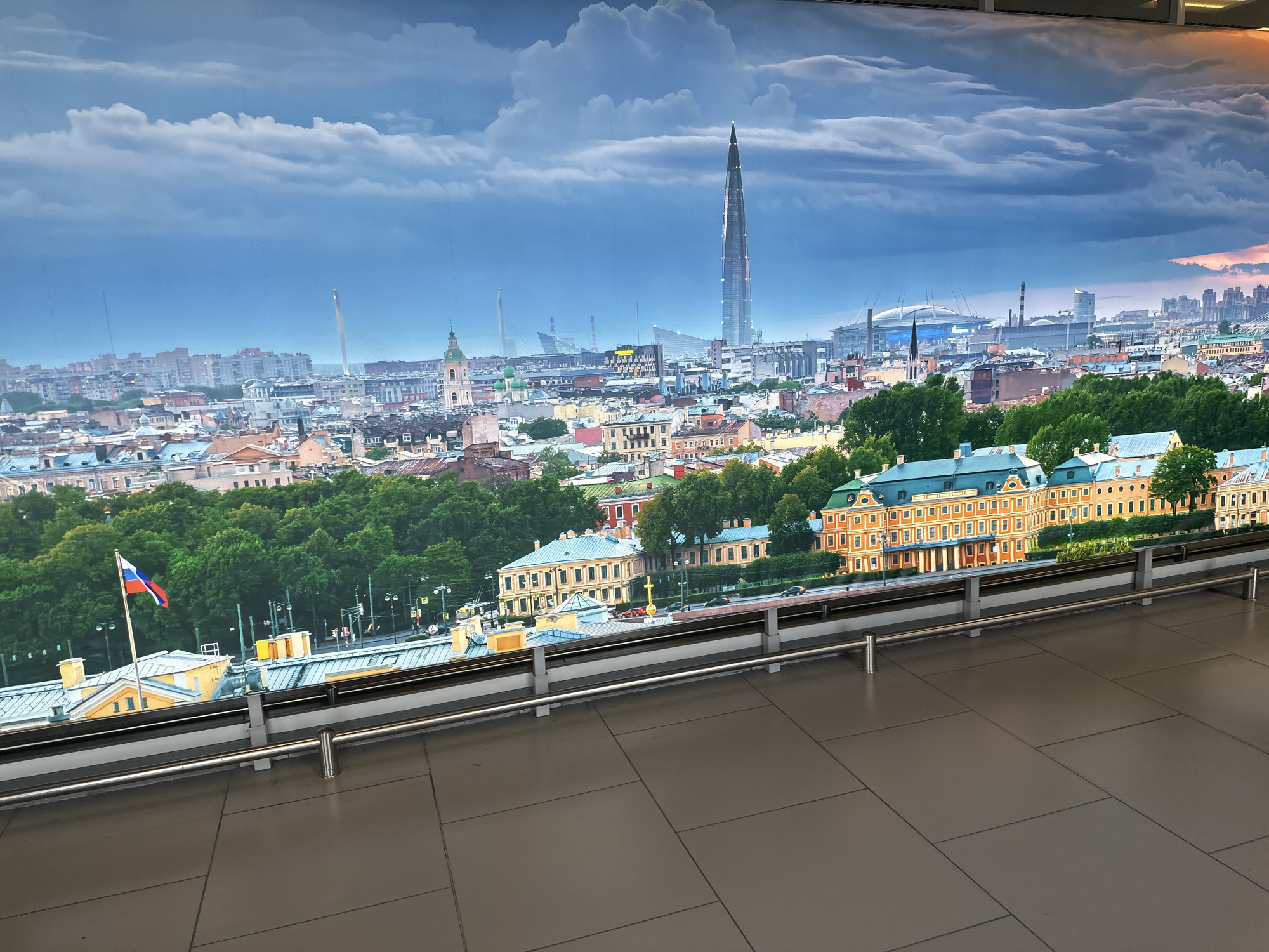 The image depicts a panoramic view of Saint Petersburg, Russia, likely a large-scale photograph or mural displayed indoors. The foreground shows a tiled floor and a stainless-steel railing. Behind the railing is the cityscape.
Visible landmarks include the Saint Petersburg Stadium, Lakhta Center skyscraper, Smolny Cathedral, and possibly sections of the Peter and Paul Fortress. A Russian flag flies atop a building in the midground. A bridge appears in the distance. The sky is filled with clouds, suggesting possible inclement weather. There are no people visible in the image.