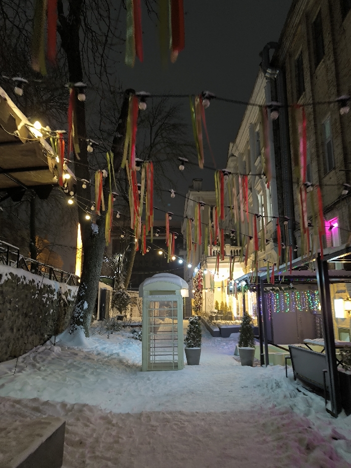 A snowy street scene at night, illuminated by strings of lights and colorful ribbons. A white telephone booth stands prominently in the center of the walkway. Buildings line the sides of the street, with some featuring decorative lighting. Snow covers the ground, trees, and surrounding surfaces, creating a winter atmosphere. Trees and bushes add to the scenery.
