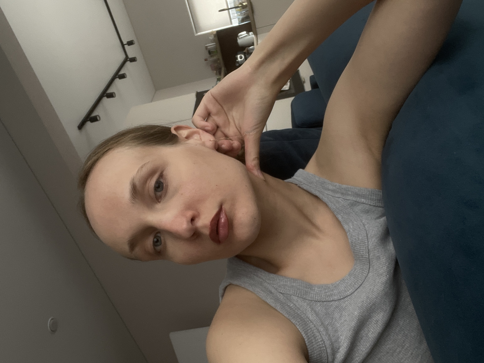 A woman with fair skin and light brown hair is sitting on a dark blue couch in an interior setting. She is wearing a gray tank top and is holding her left hand near her ear, possibly adjusting an earring. The background includes a white wall with a multi-directional light fixture, and a shelving unit with various objects. There is no definitive evidence in the image to confirm that the location is Подшивалово, Russia.