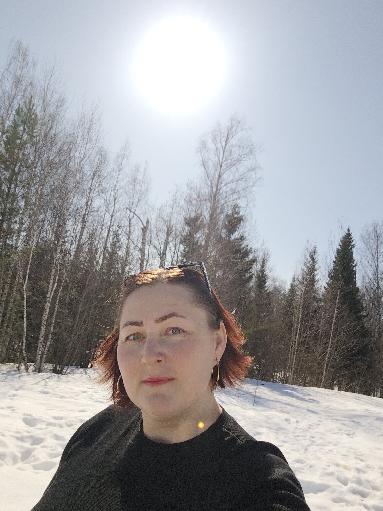 A woman is taking a selfie on a sunny day in a snowy area, likely in Марковский, Russia, given the mention of the location. She is wearing a black sweater and gold hoop earrings, with sunglasses on her head. The background consists of a forest with bare trees and evergreen trees, and the ground is covered in snow. The sun is high in the sky.