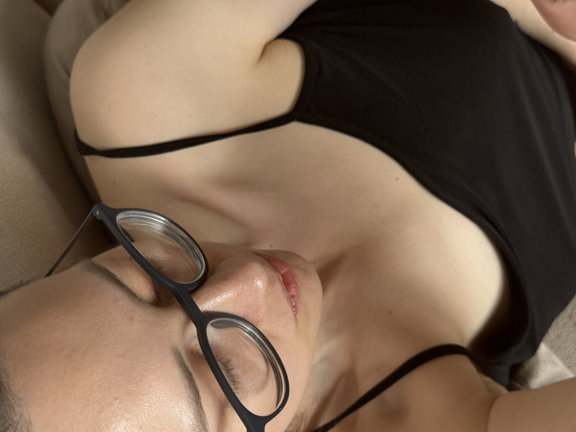 A close-up, high-angle shot depicts a person lying down, wearing glasses with dark frames. The person's skin appears light and reflective, with visible pores and eyelashes. The person is wearing a black tank top with thin straps. The background appears to be a beige or light brown fabric surface. The location cannot be determined.