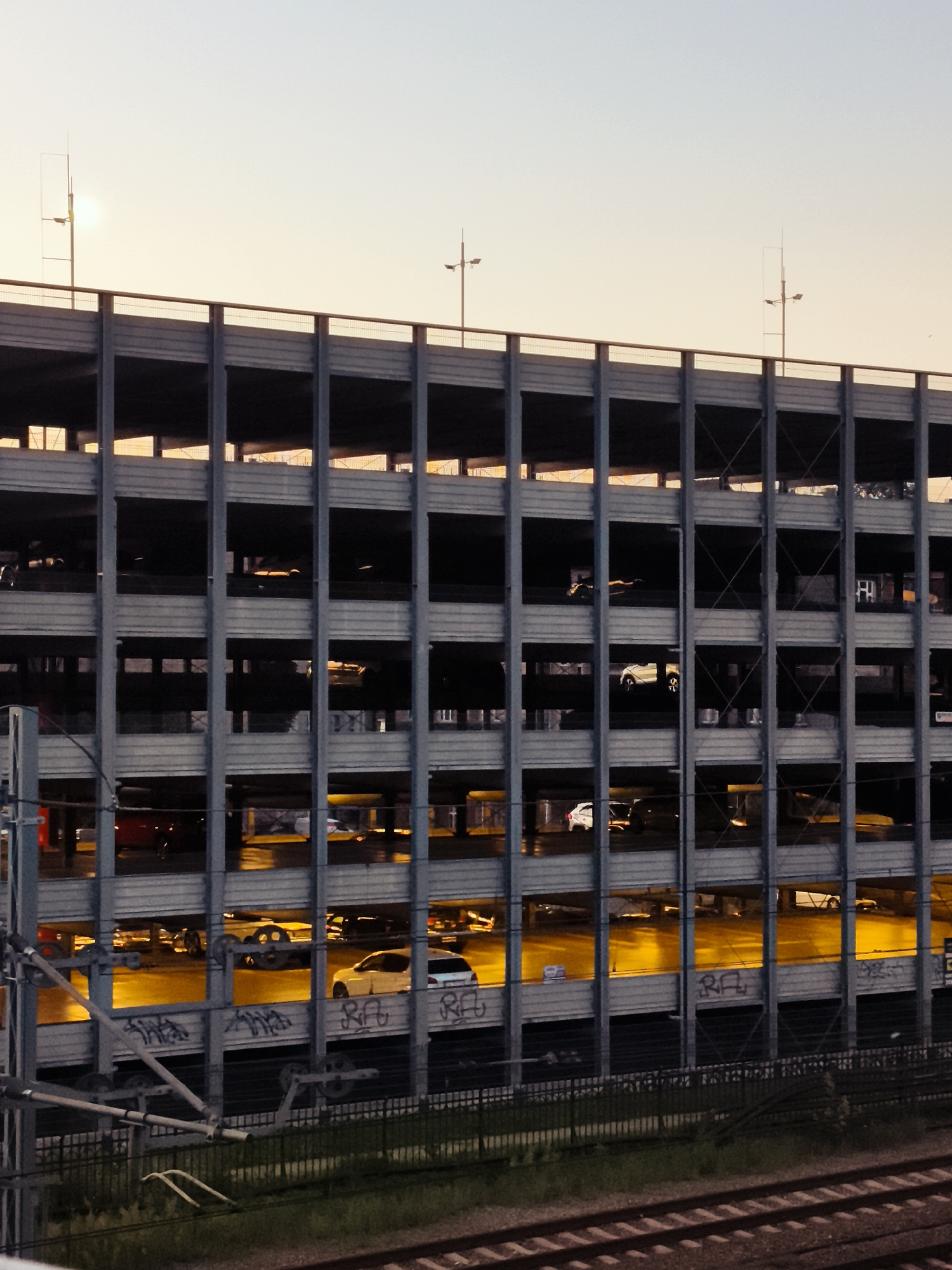 The image captures a multi-story parking garage structure with a warm, sunset sky as the backdrop. The concrete edifice is characterized by repeating horizontal and vertical beams, creating a series of open bays where several cars are parked. The lighting suggests it is late afternoon or early evening, with the setting sun casting a golden glow on the cars and the concrete surfaces. Some cars are illuminated by internal lighting, creating bright yellow reflections on the wet ground. Graffiti is visible on the lower sections of the parking garage walls. In the foreground, train tracks and a metal fence suggest the proximity of a railway line. The overall scene is urban and industrial, with the natural light of the sunset softening the otherwise stark architecture.
The parking garage appears to be located in Gdańsk, Poland, likely near the city center or a transportation hub.