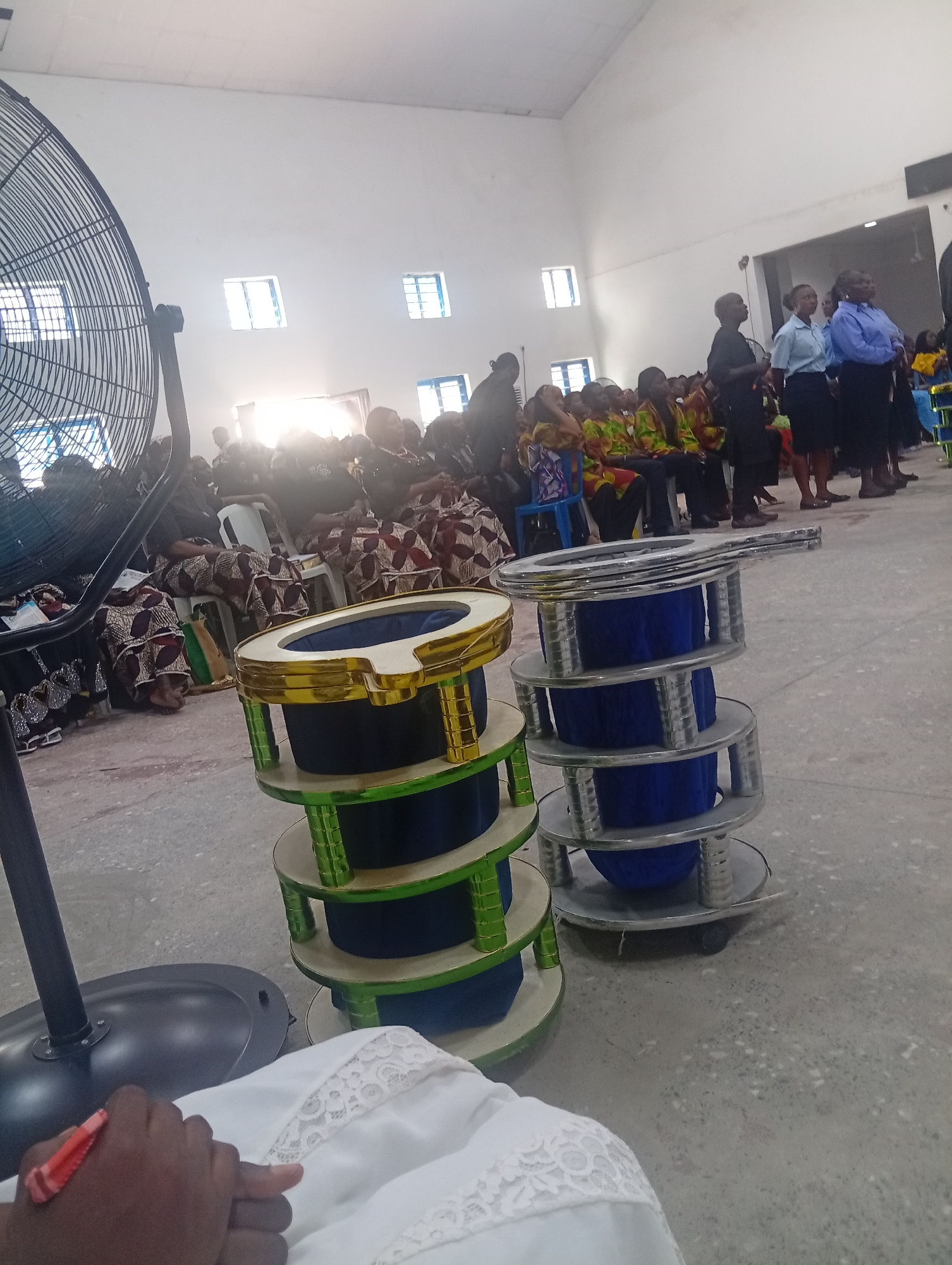 The image depicts an indoor gathering, likely a church or community hall, in Kason, Nigeria. In the foreground, a large industrial fan is positioned to the left, with its metal cage partially visible. Two tiered stands, one with green and gold accents and the other with silver accents, hold dark blue cylindrical objects, possibly decorative items or vessels. The floor appears to be concrete.
In the background, a group of people are seated and standing. Many are wearing colorful, patterned traditional African attire, while others are in more formal clothing, including shirts and skirts. Some individuals are standing and appear to be addressing or interacting with the seated group. The walls are painted white, and several windows are visible, letting in natural light, some of which is overexposed. The overall atmosphere suggests a formal event or service.
