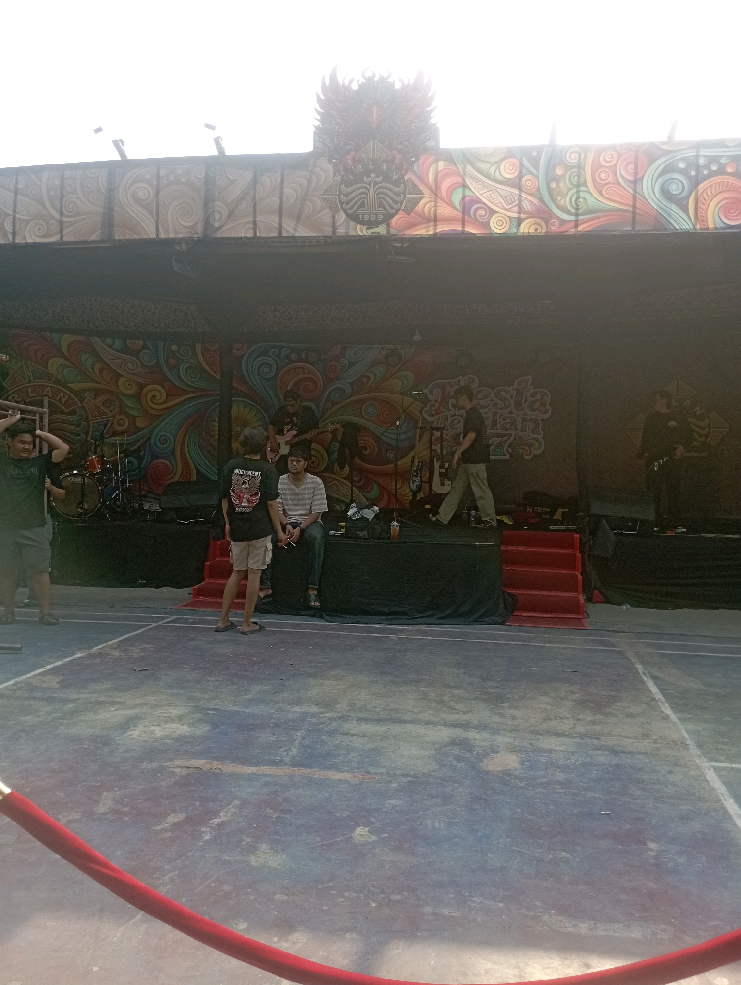 A group of young men are gathered on a concrete court in front of a stage. The stage is decorated with a large, colorful mural and features musical equipment. Two men are standing on the court, facing each other and conversing. Another man is sitting on the stage, holding a cigarette. A fourth man is standing on the stage, playing a guitar. The scene appears to be a casual outdoor gathering, possibly a concert or festival, taking place in East Jakarta, Indonesia. The ground is marked with white lines, suggesting it could be a sports court. A red rope barrier is visible in the foreground, separating the spectator area from the stage. The lighting suggests it is daytime, with the sun casting bright light on the upper part of the image. The mural behind the stage is a vibrant display of swirling patterns and geometric shapes in a variety of colors. The overall atmosphere seems relaxed and informal.
