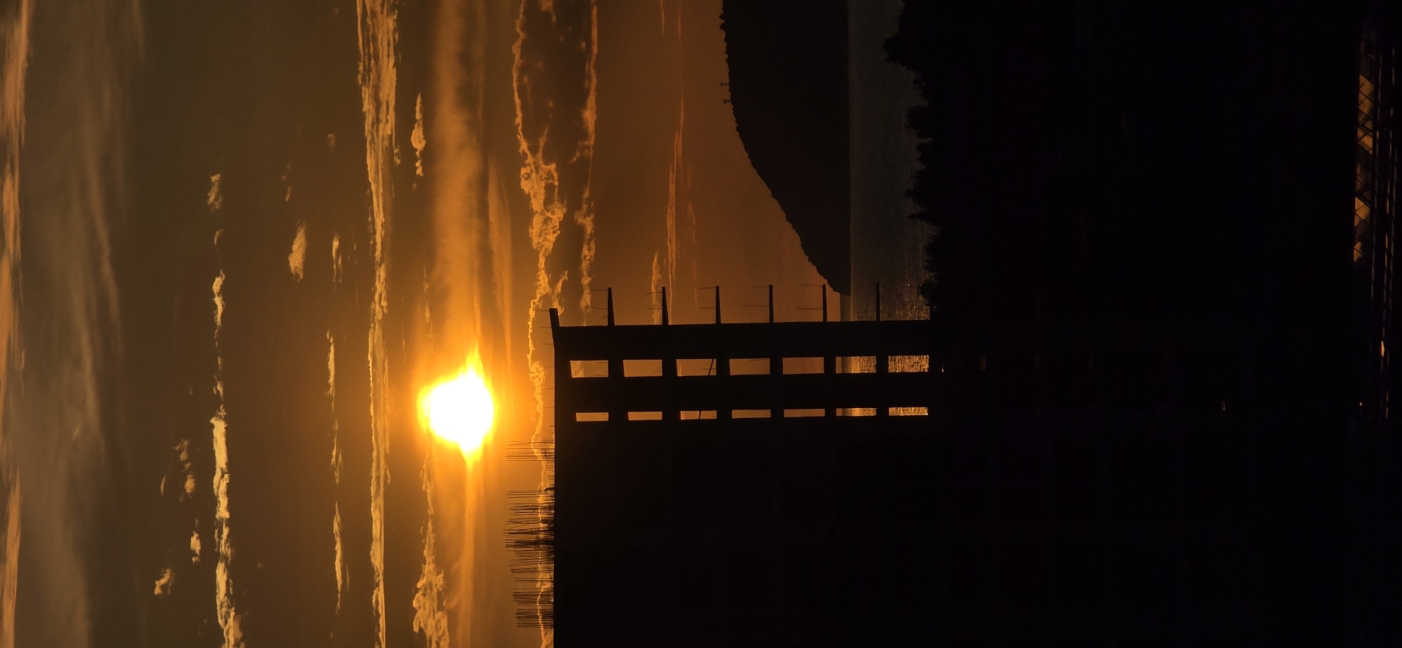 This media file captures a dramatic sunset over a body of water, likely the Adriatic Sea, in Bar, Montenegro. The sun, a bright, intense orb, is positioned low in the sky, bathing the scene in warm hues of orange, yellow, and deep red. Streaky, elongated clouds span the sky, catching the light and appearing illuminated from behind by the sun.
In the foreground, a prominent dark silhouette of a structure stretches horizontally across the lower portion of the image. This appears to be a fence, pier, or a section of a building under construction, characterized by distinct vertical posts and horizontal beams. Through the gaps in this structure, glimpses of the shimmering water are visible, reflecting the warm glow of the sky.
On the horizon, a dark, indistinct silhouette of land or mountains separates the vibrant sky from the body of water. The strong backlighting creates a stark contrast, rendering the foreground structure and distant landmass as dark profiles against the brilliant sunset. The time of day is clearly sunset, imparting a serene and awe-inspiring atmosphere to the coastal landscape. There are no people, specific activities, or visible text in the image.