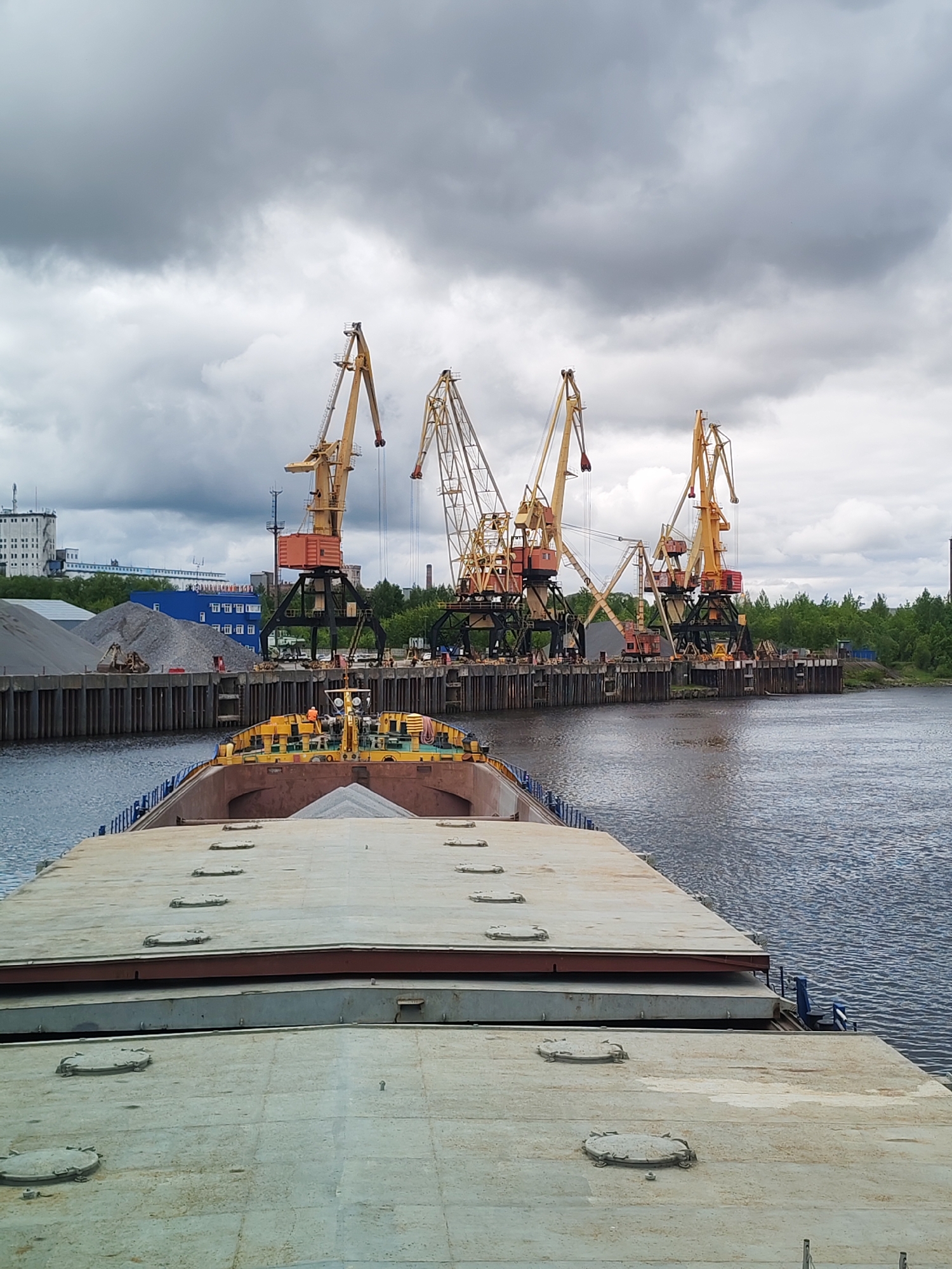 The image depicts a river scene in Rybinsk, Russia, featuring a barge in the foreground. The barge's deck is visible with multiple access hatches and a cargo of gray gravel. In the background, a port facility is present, equipped with several yellow gantry cranes positioned along a pier. Piles of gravel and construction materials lie on the pier. Buildings and green trees are also visible beyond the pier. Overcast skies cover the scene.