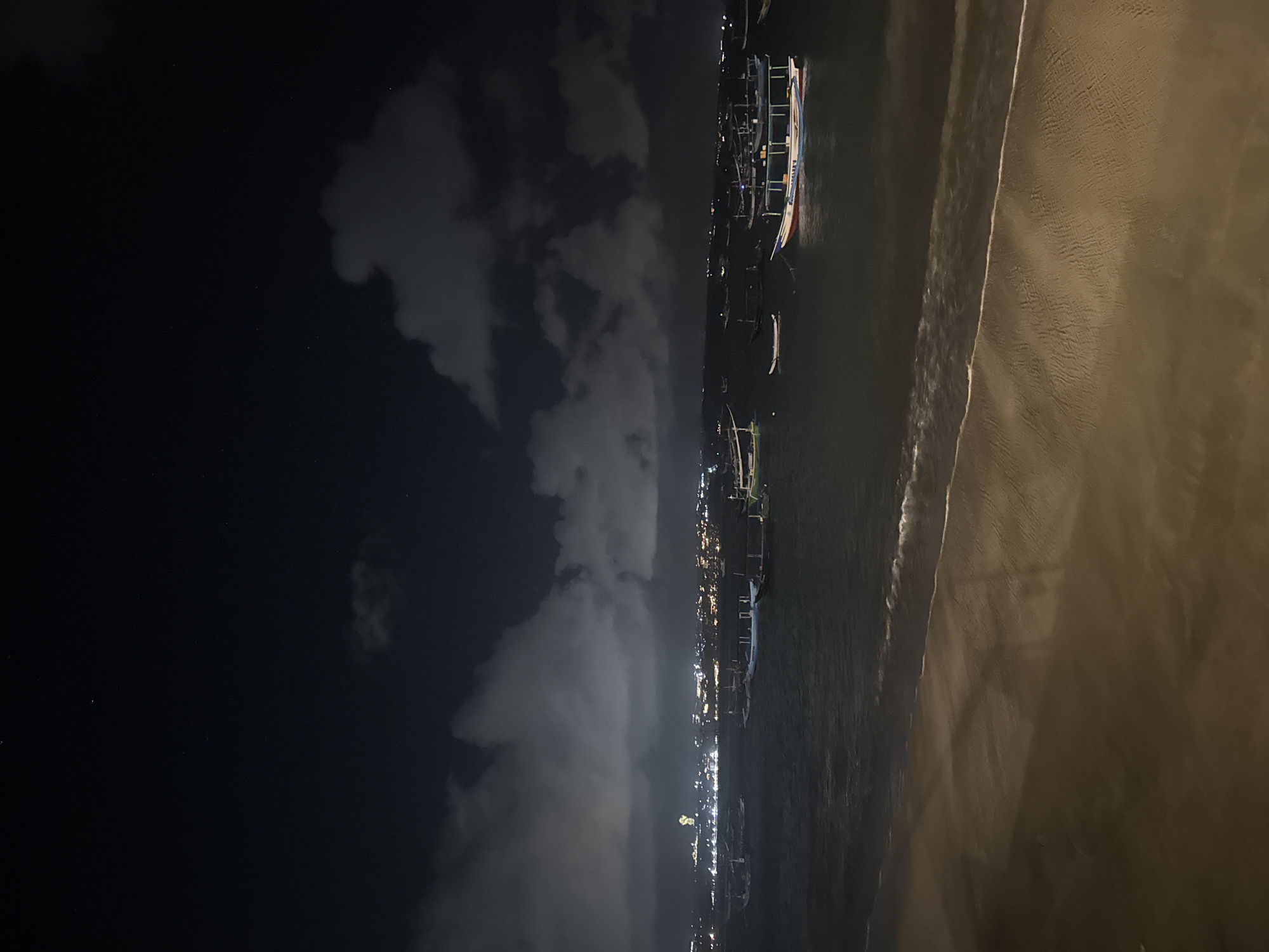 At night in Kuta, Indonesia, the scene captures the shoreline where the dark water meets the sandy beach. In the water, there are several traditional boats. The sky is dark with some visible clouds. On the shore, the beach appears wet and reflects light.