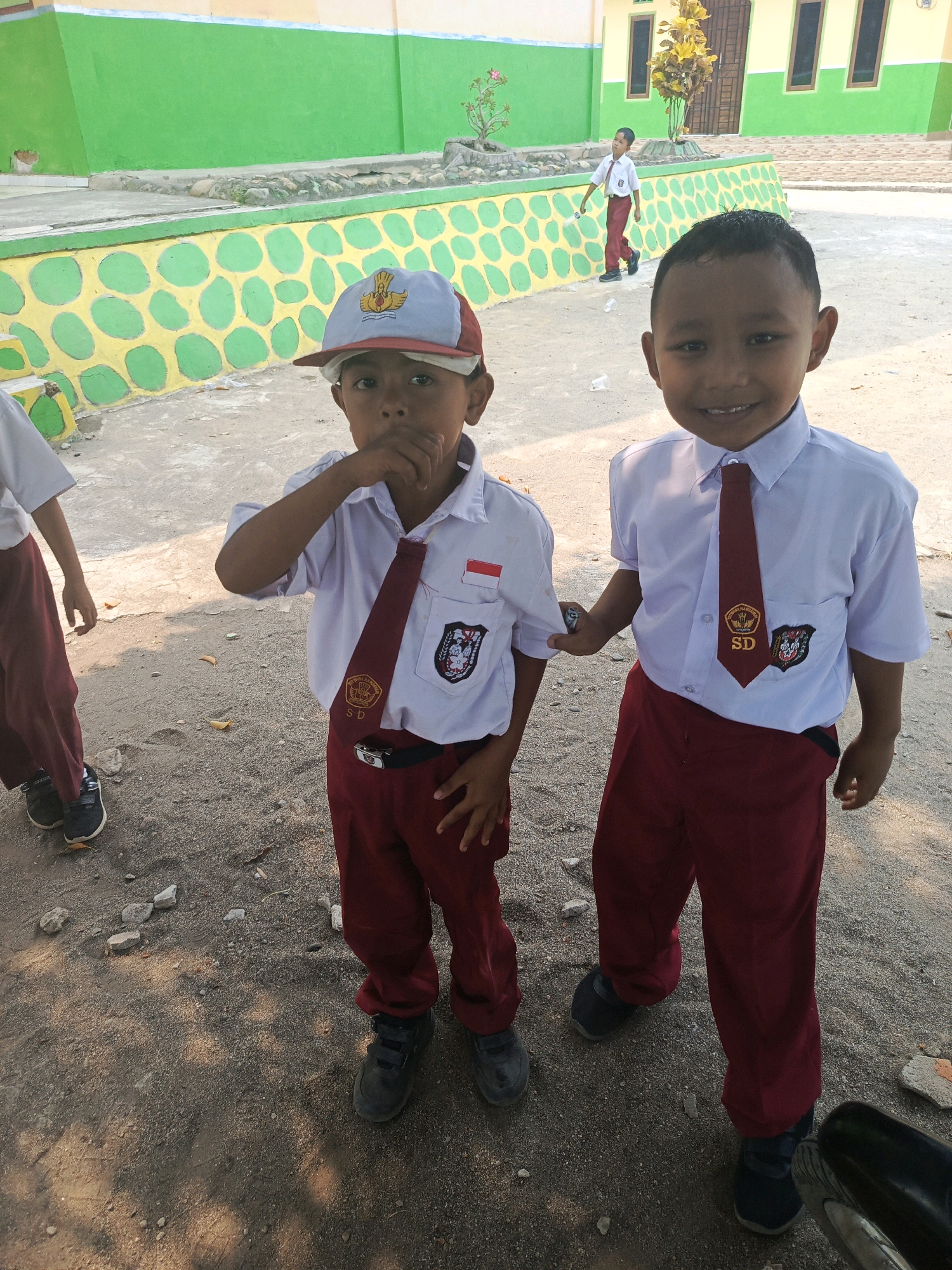 Two young boys in Indonesian school uniforms, consisting of white shirts, red ties with "SD" emblems, and maroon pants, stand on a dirt and gravel ground. The boy on the left wears a red and blue baseball cap and has his hand to his mouth. The boy on the right faces forward, smiling. Behind them, a yellow and green building with windows stands, and a smaller boy in a similar uniform walks away from the camera. The environment suggests a schoolyard in Karya Dadi, Indonesia, with a low wall decorated with yellow circles on a green background.