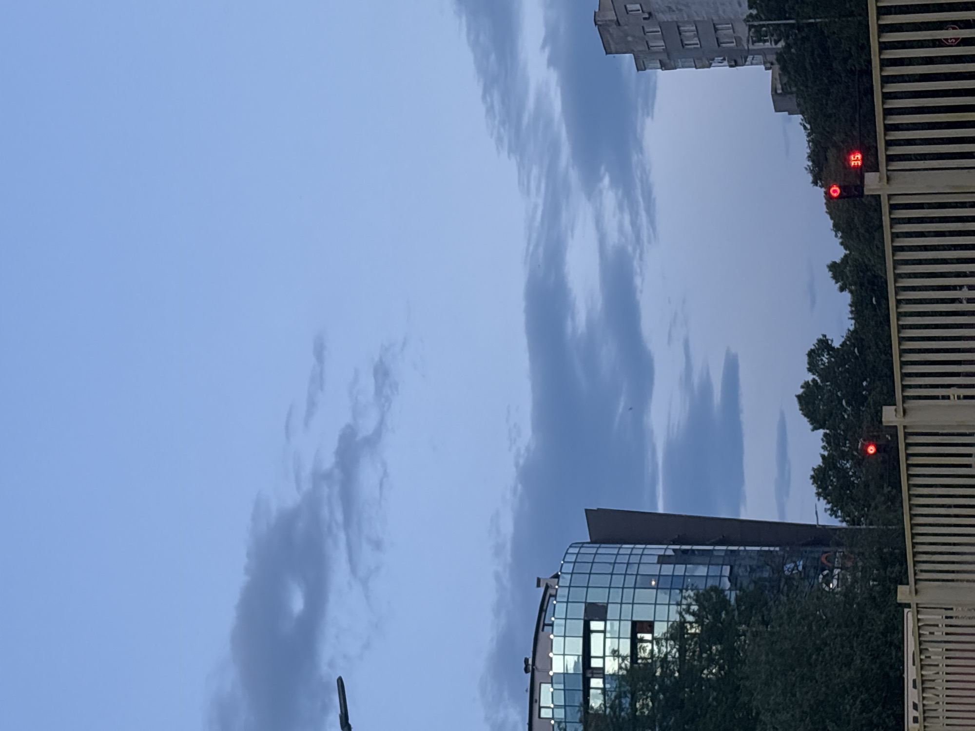 The image depicts an urban landscape under twilight conditions, characterized by a pale blue-grey sky with scattered, somewhat elongated stratocumulus clouds. In the upper left, a dark lamppost arm extends into the frame.
On the left side of the scene, a multi-story modern building with a curved, possibly cylindrical, facade is prominent. This building is primarily clad in reflective blue-tinted glass panels, which mirror the sky. Some dark architectural elements are visible to the right of this glass section. The building's roof features several small structures. Dense, dark green deciduous trees obscure the lower portion of this building.
On the right side, an older, utilitarian-style multi-story building is partially visible. It features a pale grey, possibly concrete or stucco, facade with multiple rectangular windows and balconies. Like the modern building, its lower levels are largely obscured by dark green trees.
In the mid-ground, towards the right, a black traffic light pole stands. One of its signals is illuminated red, and beneath it, a digital display shows the number "35" in red, indicating a countdown.
A light-colored, rigid barrier or fence runs across the foreground, stretching horizontally from left to right. This barrier consists of numerous vertical slats or rectangular posts, separated by gaps, topped by a continuous horizontal rail. No people or vehicles are discernible in the frame. The overall impression is a quiet street scene at dusk or dawn in the city of Unknown, Unknown.