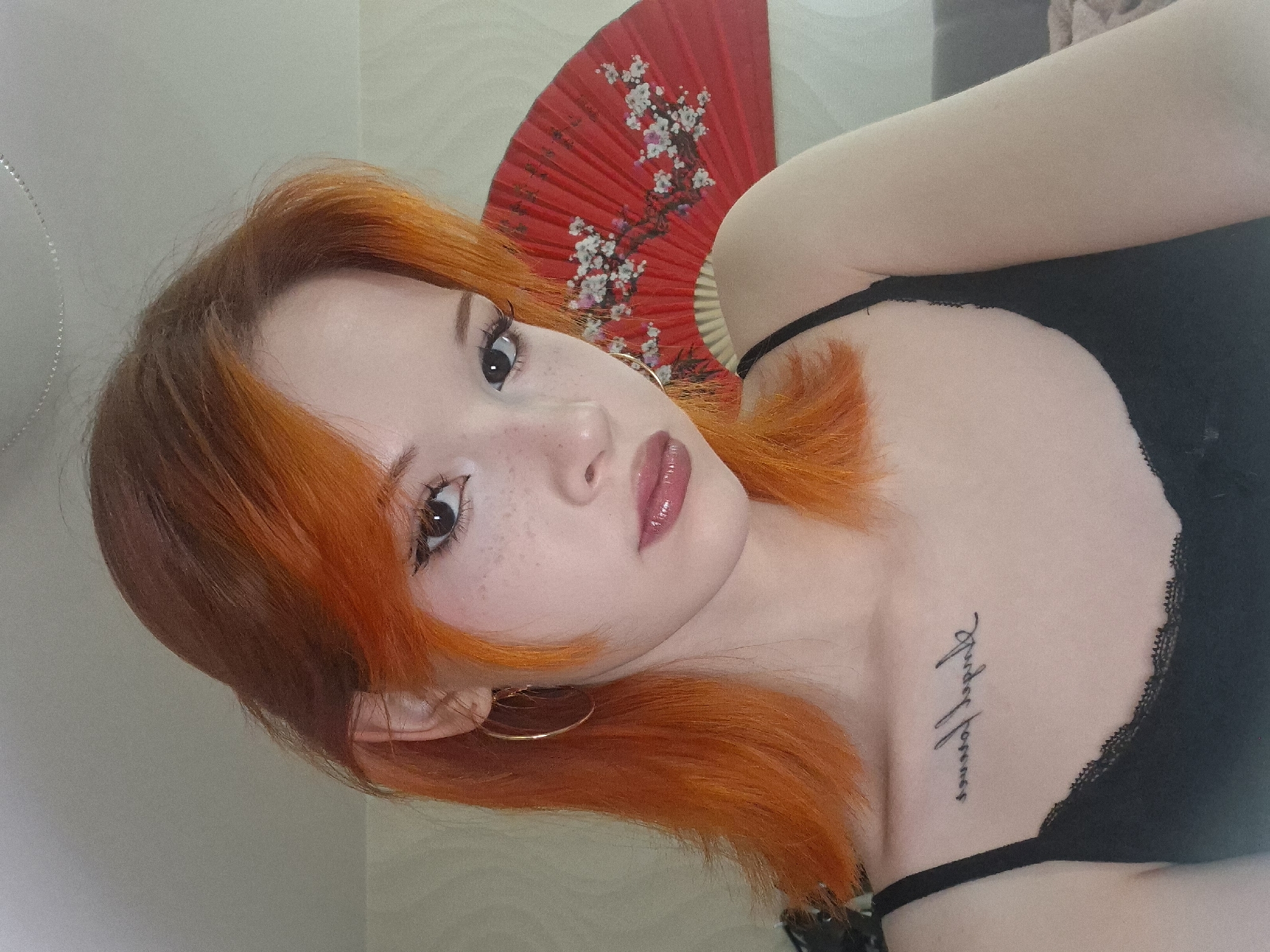 A photograph captures a young woman with orange hair, facing the camera with a direct gaze. She has freckles across her nose and cheeks and wears glossy lip makeup. Her hair is styled with bangs framing her face. She is wearing a black spaghetti-strap top with lace trim. A small tattoo is visible on her upper chest near her left shoulder. Behind her head is a red folding fan with a white floral design. She is wearing gold hoop earrings. The background is a neutral wall with a visible light fixture. There is nothing indicating the city of Chaikovsky, Russia.