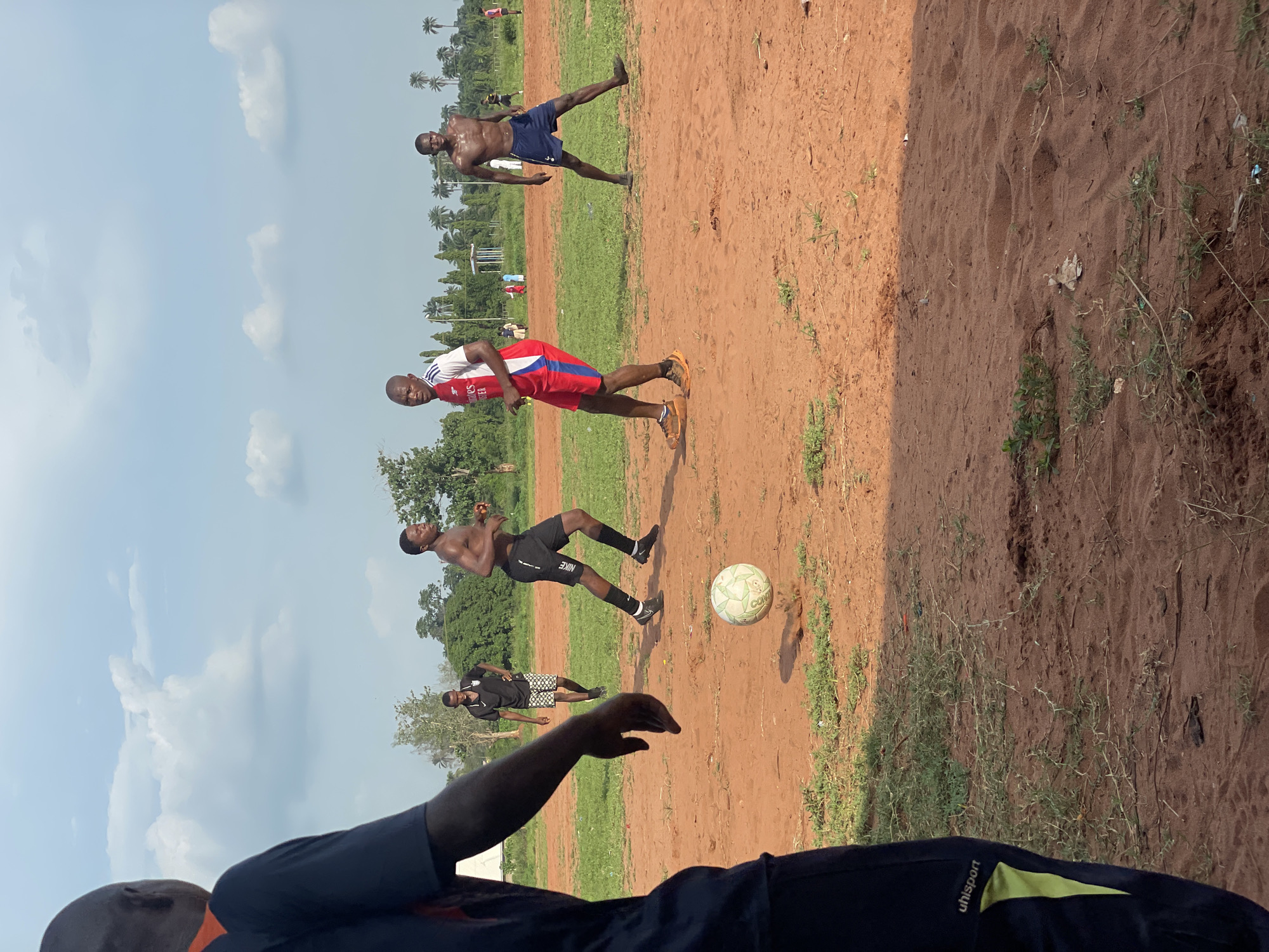 This image captures an outdoor soccer game in Ogbogbo, Nigeria, on a bright, sunny day. The scene is set on a reddish-brown dirt and sand field with patches of green grass. In the background, numerous palm trees and other dense green foliage are visible under a clear blue sky with scattered white clouds.
Several men are engaged in the game. In the immediate foreground, the back of a person wearing a dark blue shirt with "uhlsport" visible on the sleeve and green trim is partially seen, suggesting they are observing or participating from a close vantage point.
Further into the field, a man wearing a red and white athletic shirt with "ADIDAS" and "FLY EMIRATES" visible, along with black shorts and orange shoes, stands with his weight on one leg, appearing to be in motion. To his left and slightly behind him, another shirtless man in dark blue shorts stands with his legs wide apart, possibly stretching or reacting to the play.
Near the center of the frame, a man in black athletic shorts with a "NIKE" logo on the leg is captured mid-stride, seemingly running or about to kick the white soccer ball, which has "COPA" written on it, positioned to his right. In the deeper background, another player in a black and white patterned shirt and shorts is visible, along with other indistinct figures and structures suggesting a local community setting. The bright sunlight casts short shadows, indicating it is likely midday or early afternoon. The ground appears uneven, typical of an informal playing field.