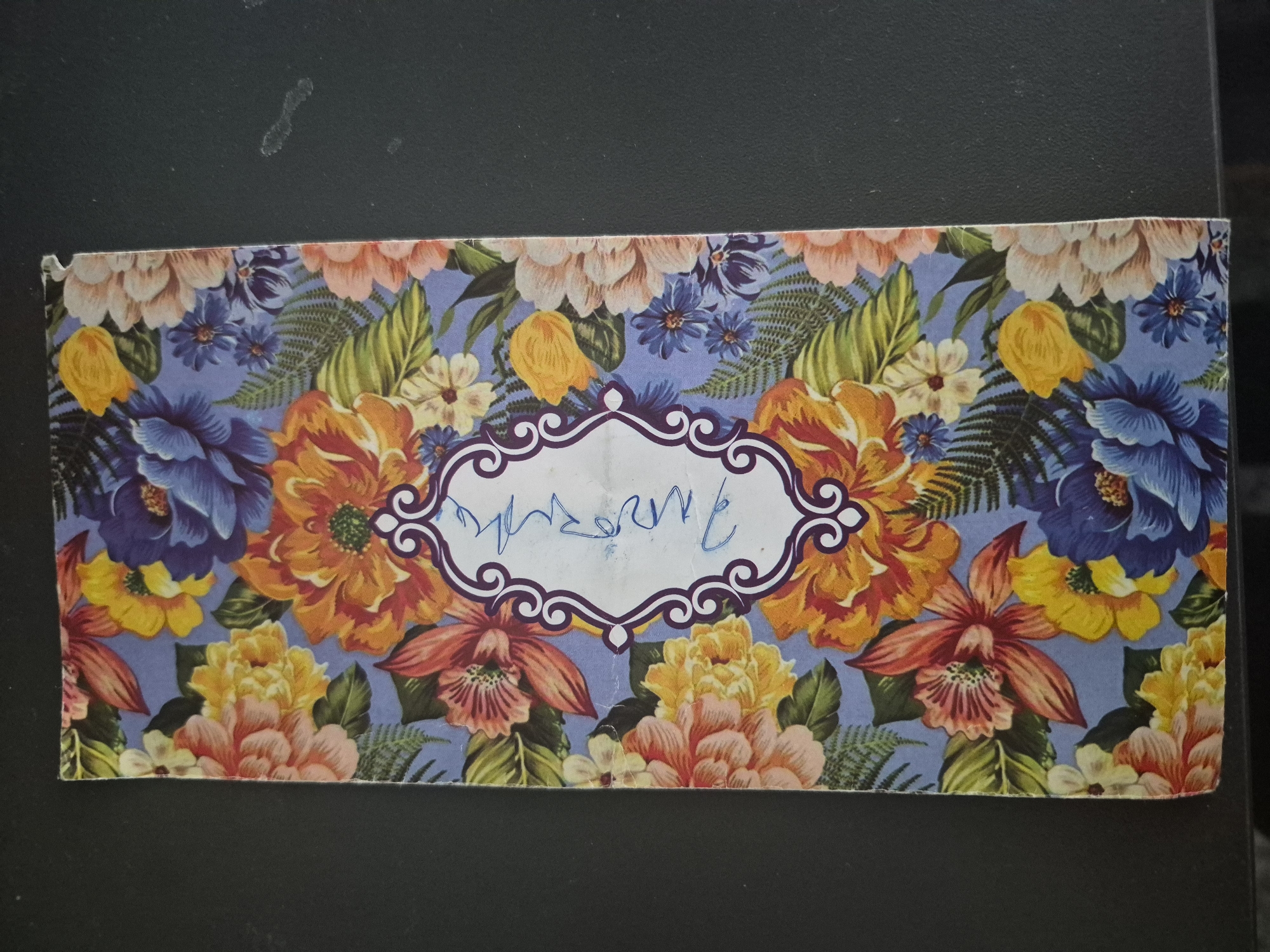 The image displays a rectangular object, likely a box or card, featuring a vibrant and dense floral pattern. The design includes prominent blossoms in shades of blue, orange, yellow, and pink, complemented by smaller white and purple flowers, and green fern-like leaves, all set against a periwinkle or light blue background.
Centered on the object is a decorative white frame with an ornate, dark purple border. Within this frame, there is illegible handwritten text in blue ink, resembling a cursive word or signature.
The object rests on a dark, flat surface, which appears to be black. There are no people or activities depicted. The edges of the object show slight wear or creasing. No specific time of day, weather, or emotions are discernible from the image. While the location context is provided as Yerevan, Armenia, there are no visual cues within the image to indicate this geographical setting.