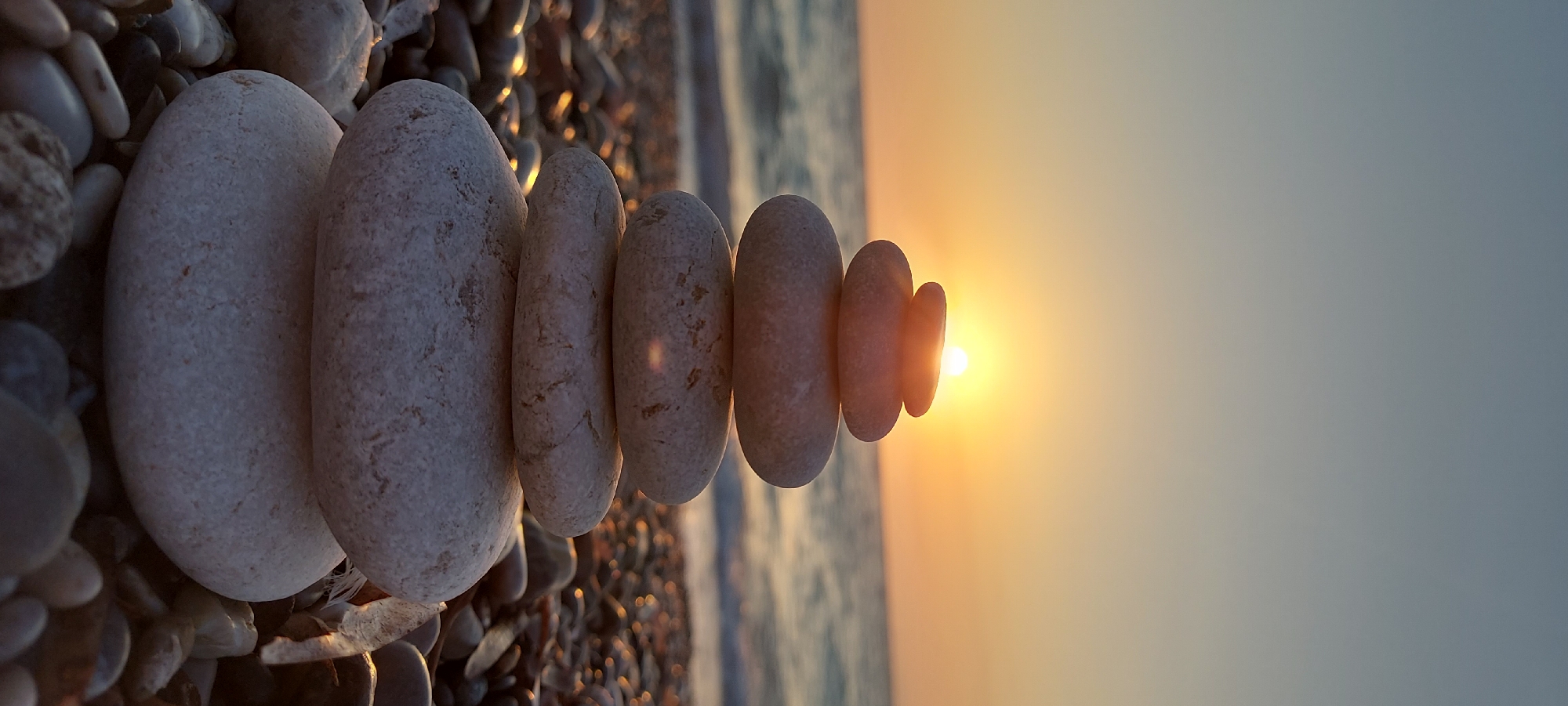 A stack of seven smooth, rounded pebbles is centered in the foreground, with the largest pebble at the bottom and the smallest at the top. The pebbles are white and grey with some texture. Behind the pebbles, a body of water, likely the sea, stretches across the frame, with gentle waves visible. The sun is setting or rising over the water, casting a warm orange and yellow glow across the sky and reflecting on the water's surface. The background is filled with pebbles and small stones on a beach. The overall scene conveys a sense of peace and balance. The image is likely from a coastal area in the Municipal Unit of Rhodes, Greece, given the characteristic pebble beaches and the clear blue waters.
