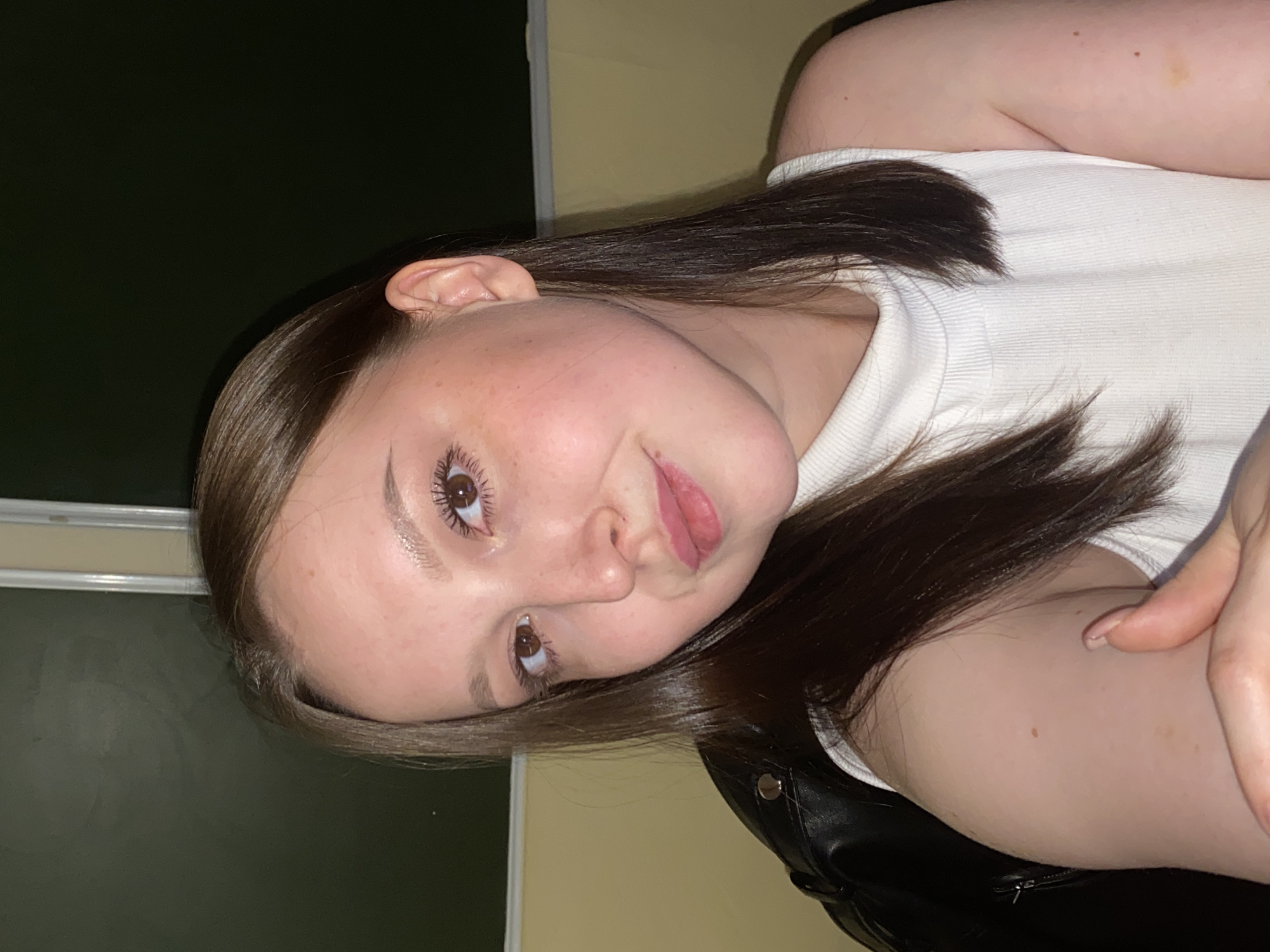 A young woman is photographed indoors, likely in Chaikovsky, Russia. Her face is the central subject, positioned slightly askew with a gentle gaze. She has fair skin, light brown eyes, and dark brown hair. She wears a white ribbed tank top and a black leather jacket with a silver snap. The background consists of a dark green wall on the left and a tan wall on the right. A window frame is visible in the upper left corner.