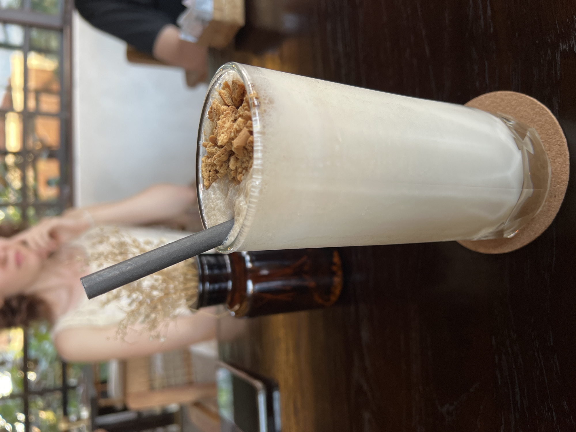 A nearly horizontal close-up shot depicts a tall glass of what appears to be a milkshake or smoothie topped with crumbled cookies, positioned on a dark wooden table with a brown coaster underneath. A grey straw sticks out of the glass. To the left and partially obscured is a small brown bottle, possibly containing syrup or flavoring. In the background, out of focus, a fair-skinned person wearing a light-colored garment is visible, along with the blurred impression of a window or outdoor scenery, suggesting a restaurant or cafe setting. The photograph appears to have been taken in the Ubud District, Indonesia.