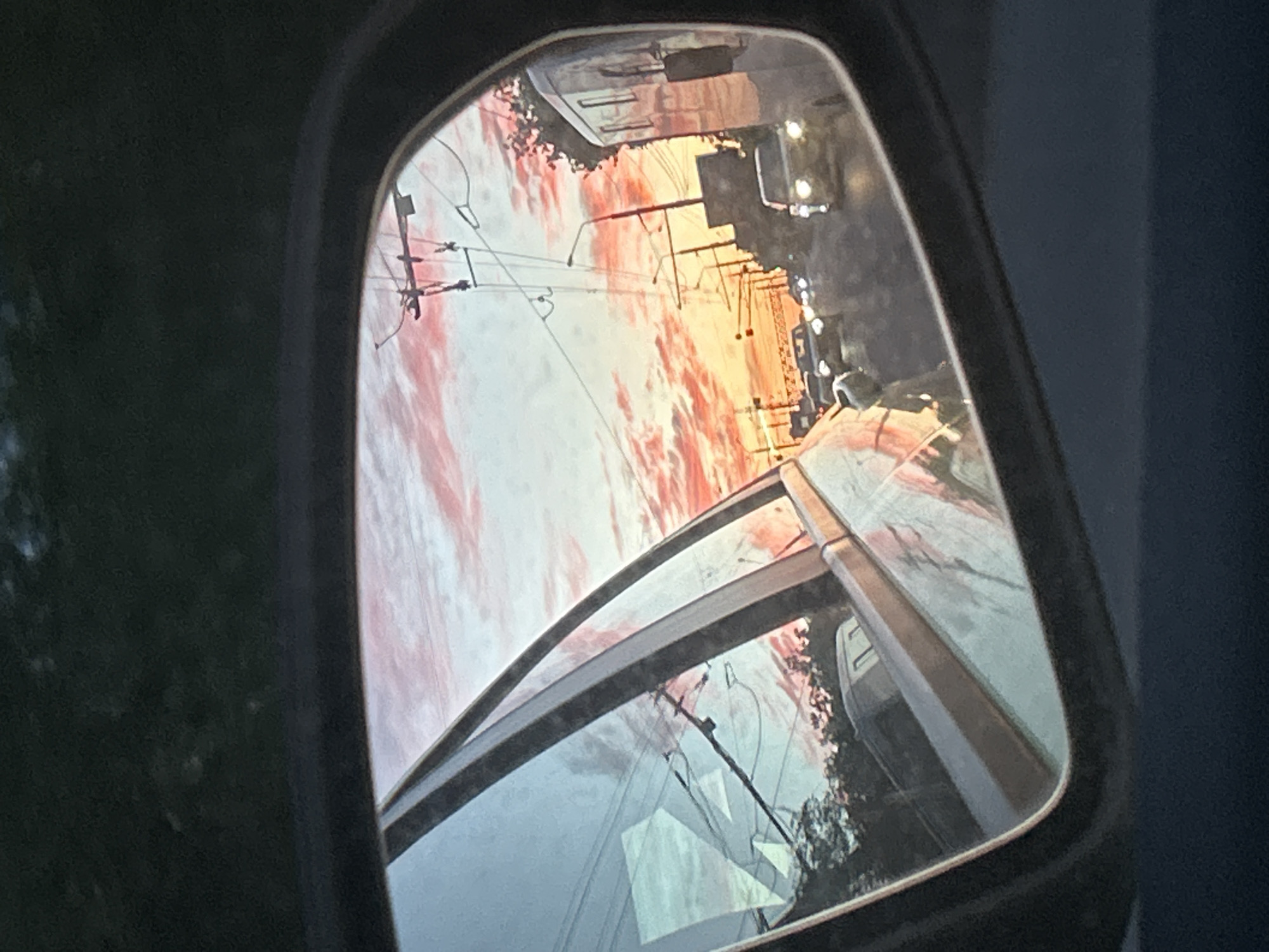 The image shows a car's side mirror reflecting a sunset view of Тимофеевка, Russia. Power lines and poles run along a street. Several vehicles are visible. The sunset creates a red and orange sky with clouds.