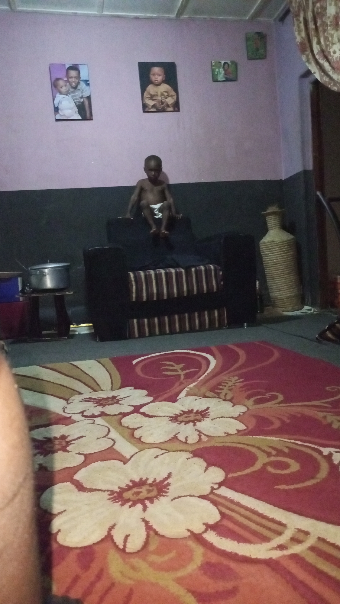The image shows a domestic scene in Sabon Tasha, Nigeria. A young child wearing a diaper sits on the back of a dark upholstered armchair, which is positioned against a wall that is painted dark on the bottom half and light purple on the top half. Above the chair are three framed pictures hanging on the wall. To the left of the chair, a small side table supports a cooking pot. To the right of the chair is a tall, decorative vase. In the foreground, a red patterned rug with large floral designs covers the floor. Part of a person's arm is visible in the bottom left corner of the image.