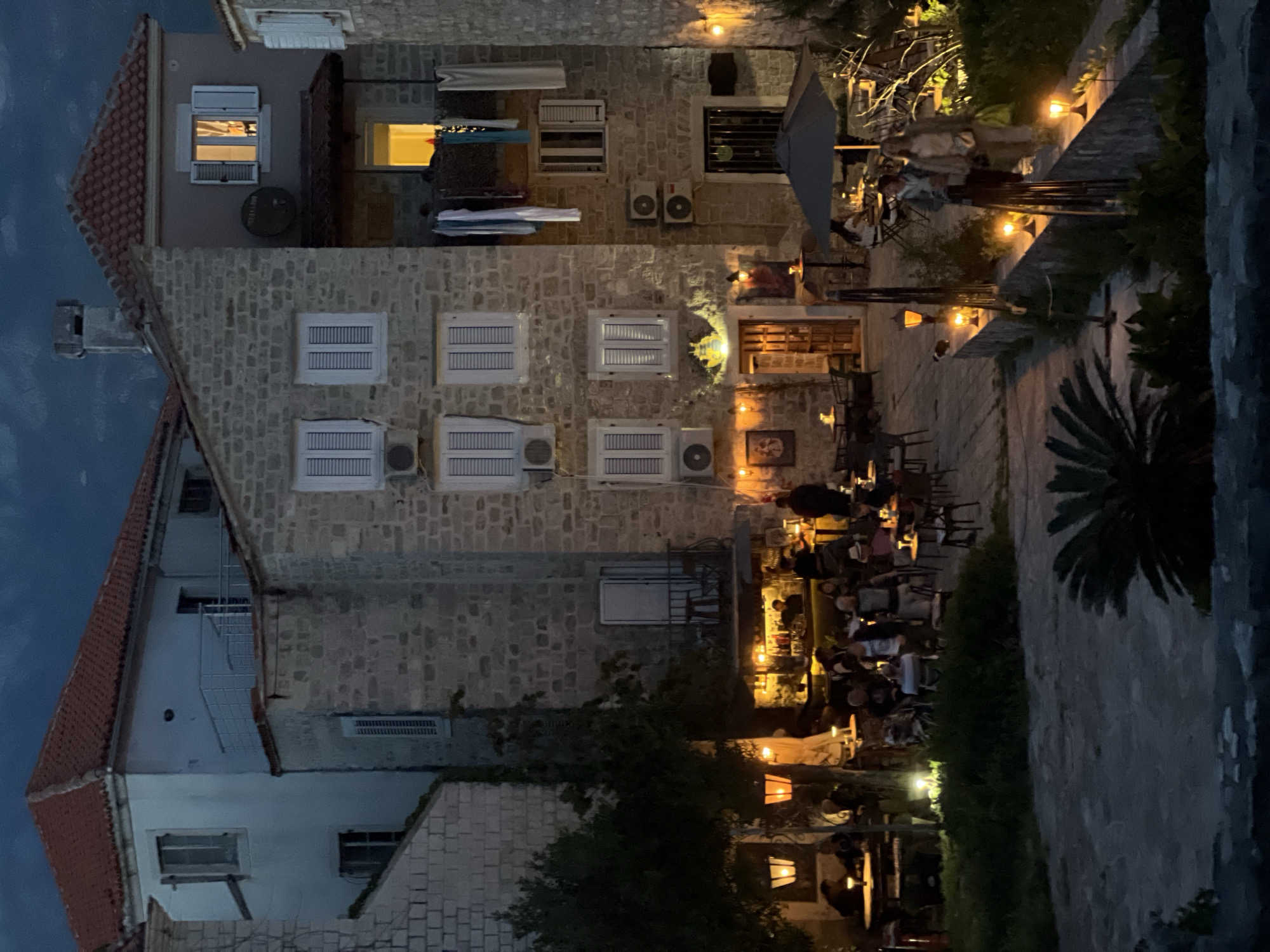 The image depicts an evening scene in Budva, Montenegro, characterized by its old stone buildings and a lively outdoor dining area.
**Setting:** The scene is set in a narrow outdoor alley, bordered by the aged stone facades of buildings with red tile roofs. The alleyway is paved and dimly lit by overhead lamps and the glow emanating from the restaurants.
**Objects:** The buildings exhibit architectural details such as windows, air conditioning units, and satellite dishes attached to the walls. The outdoor dining area features tables and chairs set up along the alley, with umbrellas providing partial cover. Potted plants and greenery are strategically placed to enhance the ambiance.
**People:** Numerous patrons are seated at the tables, dining and engaging in conversation. Their specific actions are not clear due to the distance and dim lighting, but it can be assumed they are primarily eating, drinking, and socializing.
**Overall Impression:** The image suggests a vibrant and inviting atmosphere in the historic part of Budva, Montenegro, with the outdoor dining scene serving as a focal point. The combination of old-world charm and modern hospitality is apparent.