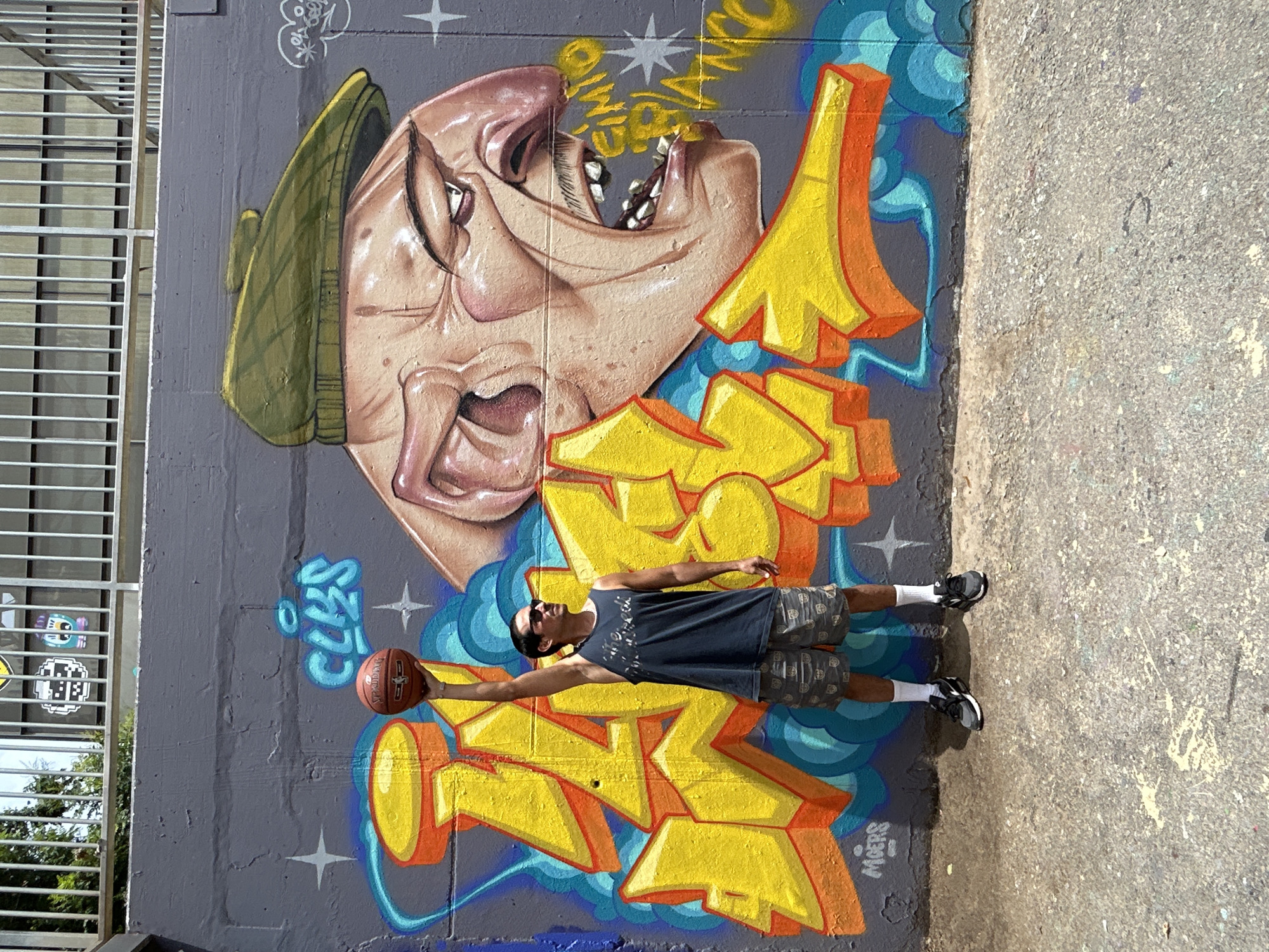 A man stands holding a basketball above his head in front of a large graffiti mural. The mural depicts a cartoonish caricature of a man's face wearing a green flat cap, with his mouth open. Behind the face, large, stylized yellow letters are painted, along with blue cloud-like shapes and white starbursts. The man is wearing a dark tank top, camouflage shorts, white socks, and black sneakers. He is also wearing sunglasses. The graffiti is on a grey concrete wall, and the ground in front of it is also concrete. A metal railing is visible above the wall, with some sticker art adhered to it. The scene appears to be outdoors, possibly in an urban environment. The graffiti itself contains some writing in yellow, with one part appearing to say "MIGERS" in white. The other yellow text is harder to decipher but may include "CIMIO" and "BLANCO". The man's posture suggests he is posing for a photo.