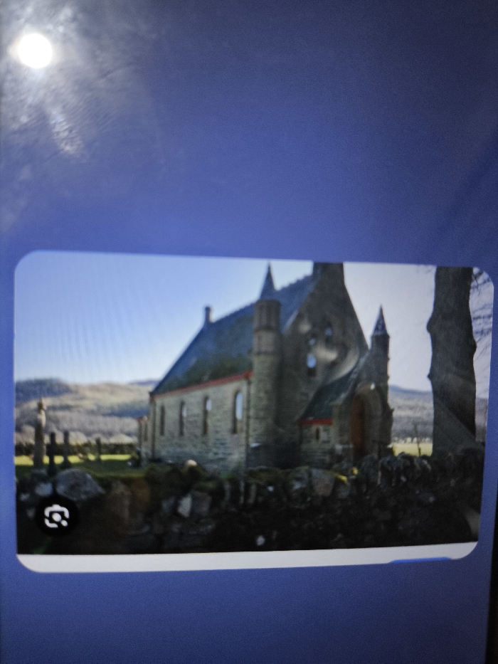 The image displays a photograph of a digital screen exhibiting an outdoor scene. The central subject is a stone church building, located in the city of Nicosia, Northern Cyprus / Cyprus. This structure features light-colored stone masonry, a dark pitched roof, and multiple pointed turrets and spires. Several vertical rectangular windows, accented with reddish-orange trim, are visible along its side. To the left foreground, a graveyard is indicated by multiple weathered stone crosses and markers, partially obscured by a low accumulation of rocks or a dry stone wall. The immediate foreground also shows green grass. The background consists of rolling hills with sparse vegetation under a clear, bright blue sky. On the far right, the trunks of two large, bare trees are present. No individuals are depicted. The photograph of the screen itself shows a bright, circular light reflection in the upper-left quadrant and a small visual search icon in the lower-left corner of the displayed image frame.